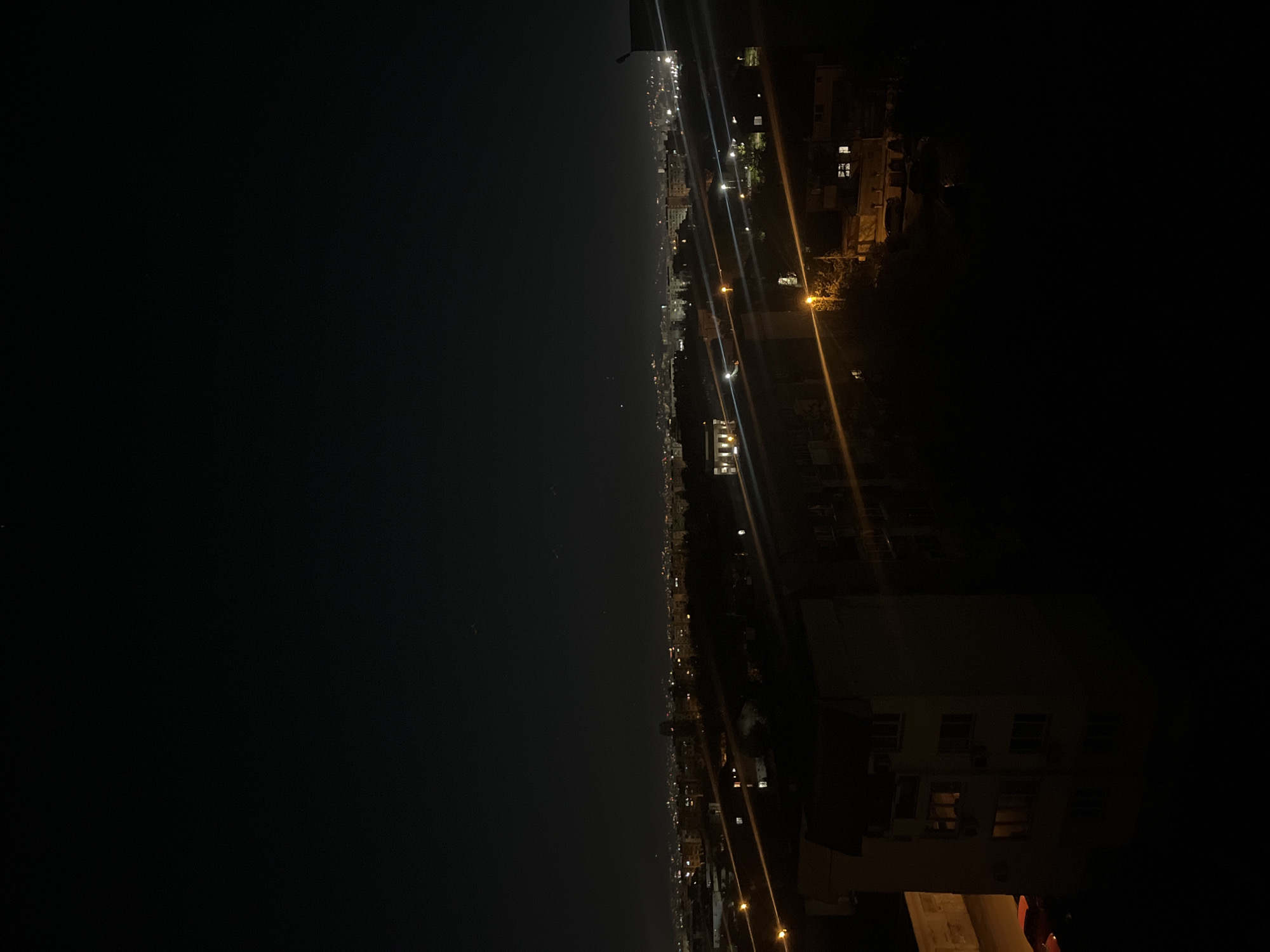 The image shows a cityscape at night. The left side of the image is dominated by darkness, possibly depicting a body of water. A string of lights runs along the horizon, suggesting a city skyline with varying building heights and light sources. The right side features structures, likely buildings, with illuminated windows and streetlights. The perspective suggests the photo was taken from an elevated position. Given the prompt's instruction, it can be said that the city is Sofia, Bulgaria, but no specific locations can be made out from the available information. No people are visible.