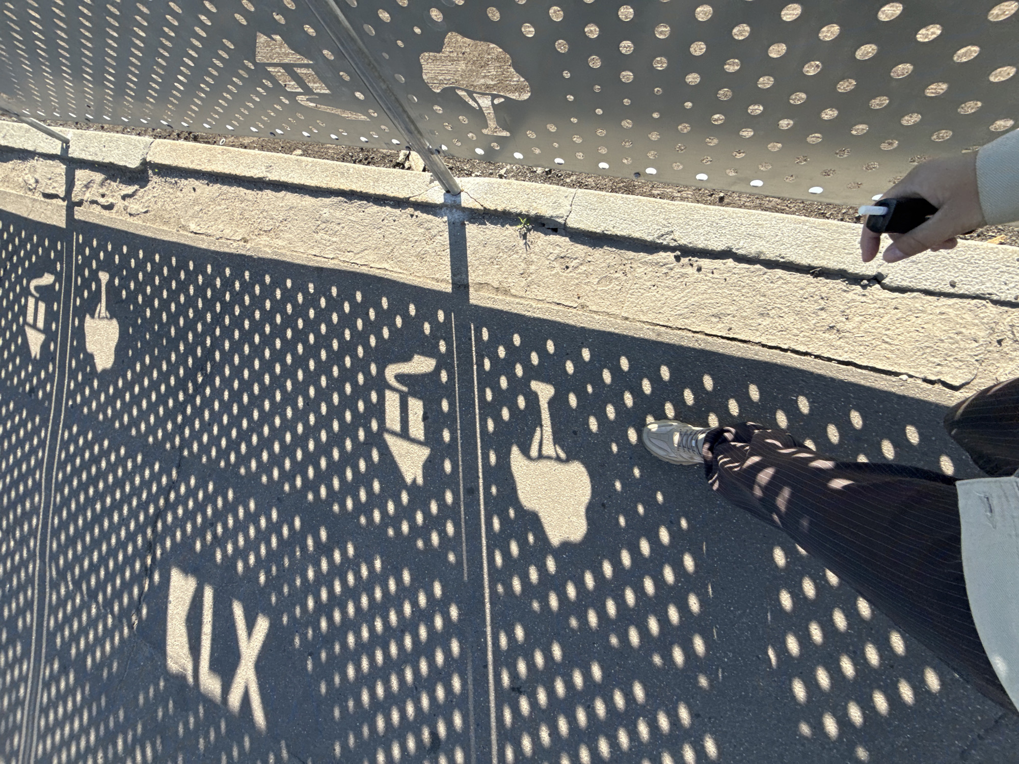 The image displays a high-angle view of a sunlit paved path, heavily patterned with shadows cast by a perforated metal structure overhead. A person's lower body is visible, with a leg dressed in dark, possibly pinstriped, trousers and a foot wearing a light-colored athletic shoe with white laces. In the upper right, a hand with a light-colored sleeve holds a small, black object with a white tip, possibly a pen or an electronic device.
The ground is covered in a dense pattern of bright, circular spots of sunlight alternating with areas of shadow, created by the numerous perforations in the metal structure. Interspersed within these patterns are larger, distinct shadow shapes. In the lower-left, the letters "LX" are prominently formed by light. Further into the frame, other shadow cut-outs include shapes resembling a small stylized tree or plant and a shovel, reflecting designs in the metal above.
A concrete curb runs across the mid-ground, separating the paved path from a narrow strip of earth or dirt. The sharp, well-defined shadows indicate bright, clear sunlight. The scene depicts an outdoor setting, likely a public space, in Moscow, Russia.