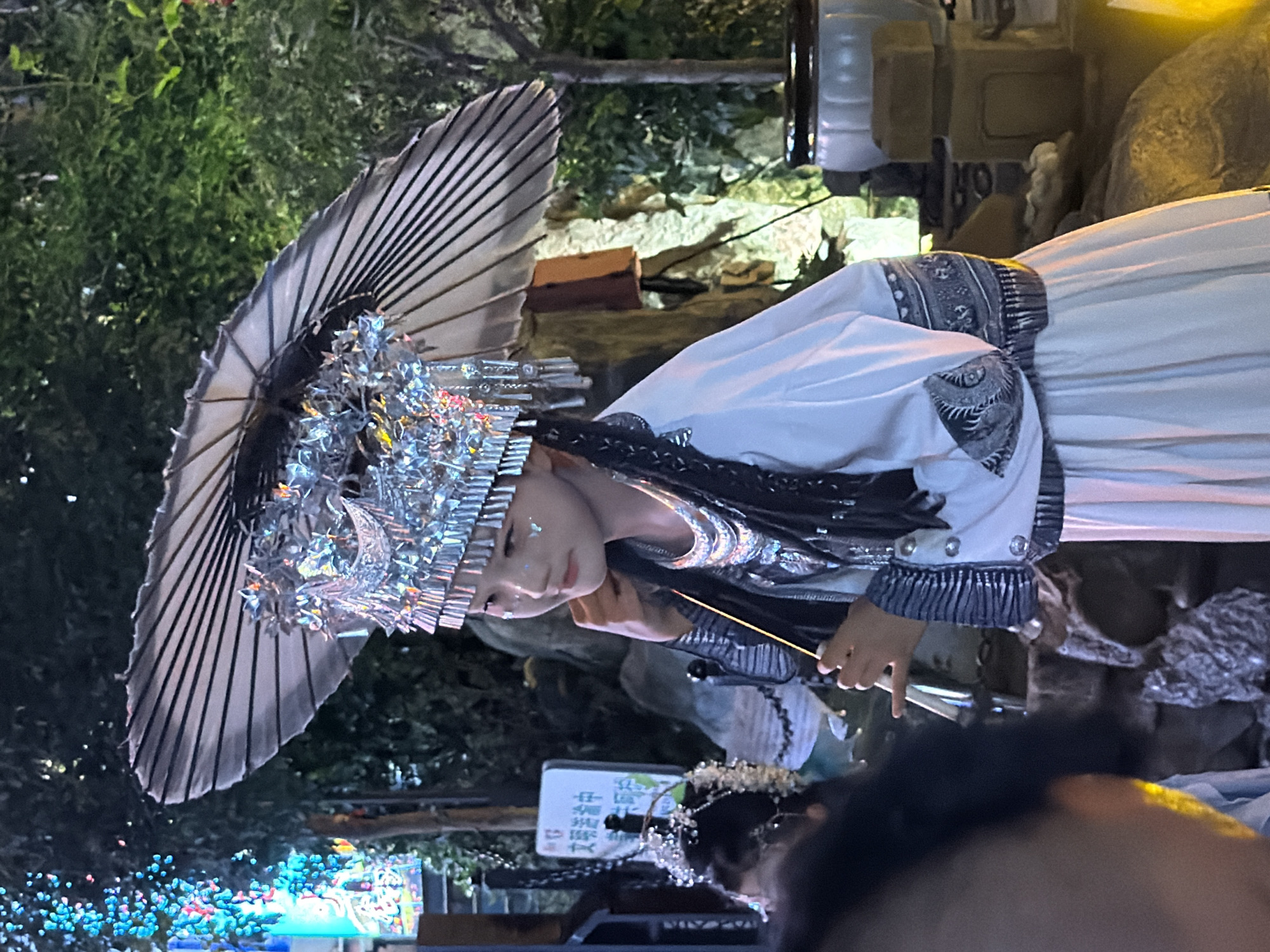A young person in traditional Yuzhong District, China attire is standing outdoors at night, holding a decorative parasol. The person is wearing a white, flowing dress with intricate blue and silver patterns on the sleeves and hem. A prominent, ornate silver headdress adorned with tassels and reflective elements sits atop their head, partially obscuring the parasol's handle. The person's face is turned to the side, with a subtle smile and delicate butterfly-shaped glitter on their cheek. They are holding a thin, light-colored stick vertically in their right hand. In the background, lush green foliage is visible, along with some illuminated decorative lights that cast a colorful glow. There are also elements of what appears to be a rocky landscape and traditional Chinese architectural features. The overall scene suggests a cultural performance or celebration taking place in Yuzhong District.