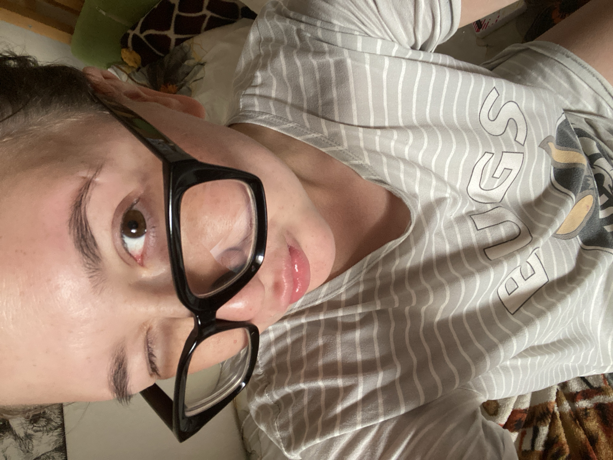 A woman with brown eyes and dark hair is wearing large black-rimmed glasses. She is winking and puckering her lips. She is wearing a grey and white striped t-shirt that has the word "UGS" visible on it, as well as part of an image of a cartoon dog. The background appears to be a bedroom with a giraffe-patterned pillow and a green pillow. This image was taken indoors.