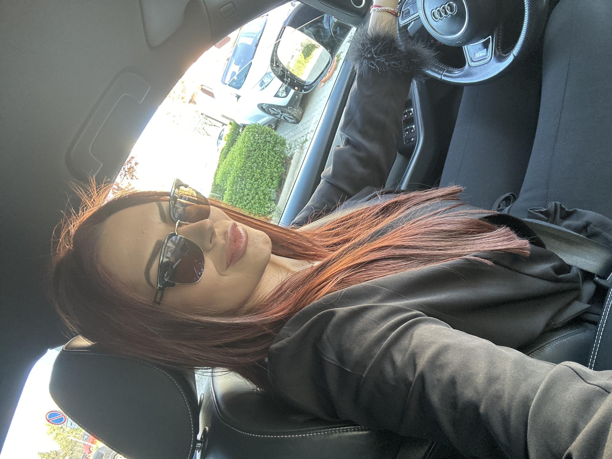 A woman with reddish-brown hair, wearing sunglasses and a black jacket, is in the driver's seat of an Audi. She is buckled up with the seatbelt visible. The car's steering wheel is visible, and a fuzzy accessory is attached. Outside the car, a street scene shows a white car parked and green bushes. Based on the traffic sign visible in the lower-left corner, the location is identified as Sofia, Bulgaria.