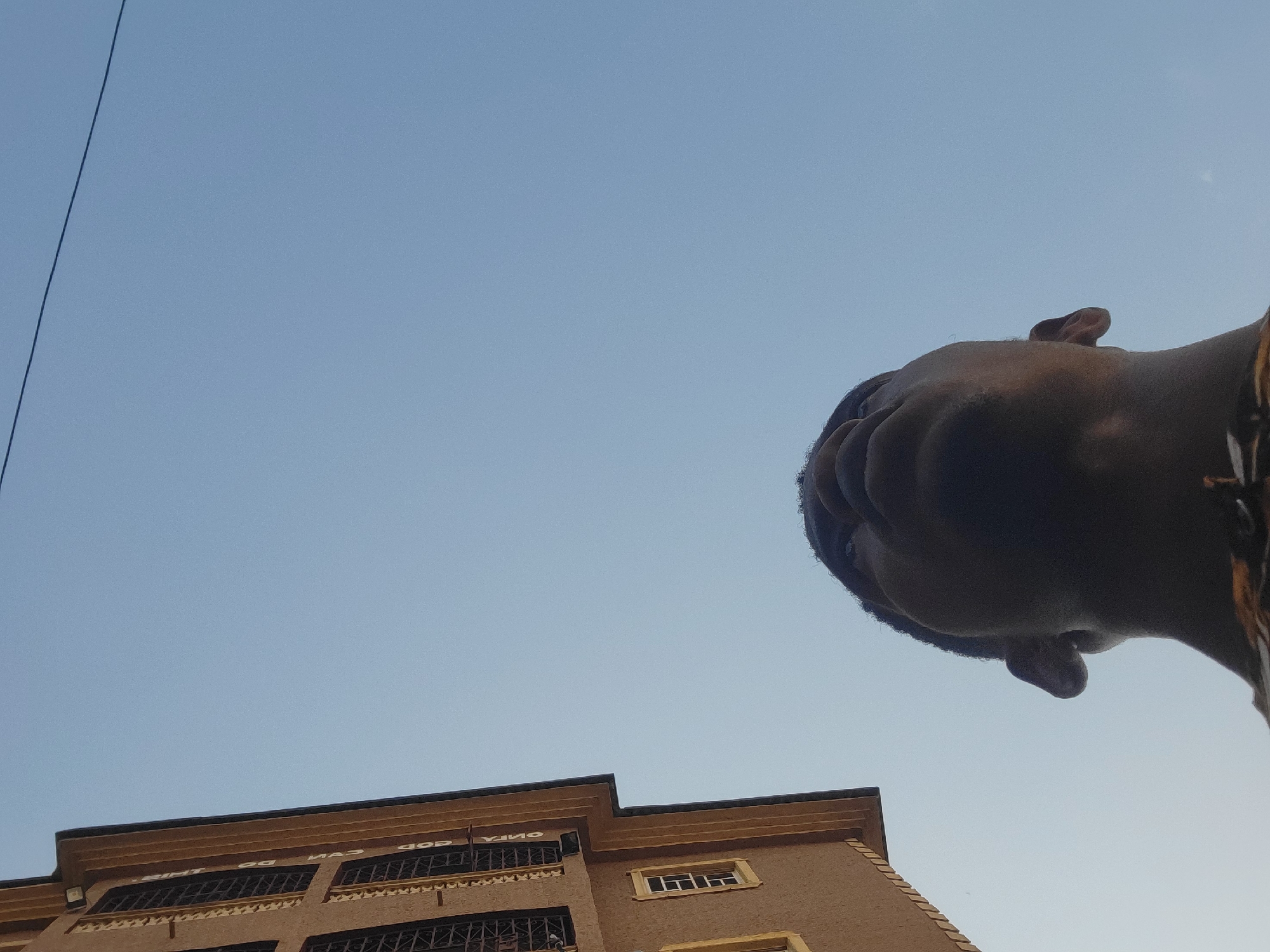 A low-angle shot captures a clear blue sky above a building and a person's head. On the left side of the frame, a multi-story building with cream-colored walls and dark, barred windows is visible. The building appears to be in the city of Onitsha, Nigeria, suggested by the style of architecture and the context provided. The person in the foreground is a Black male, whose head and upper torso are the primary focus. He is looking upwards, with his face partially obscured by the camera's perspective. The background is dominated by the expanse of the sky, with a thin power line stretching across the upper portion of the image. The overall impression is one of looking up towards the sky, with urban surroundings present but secondary to the sky and the individual.