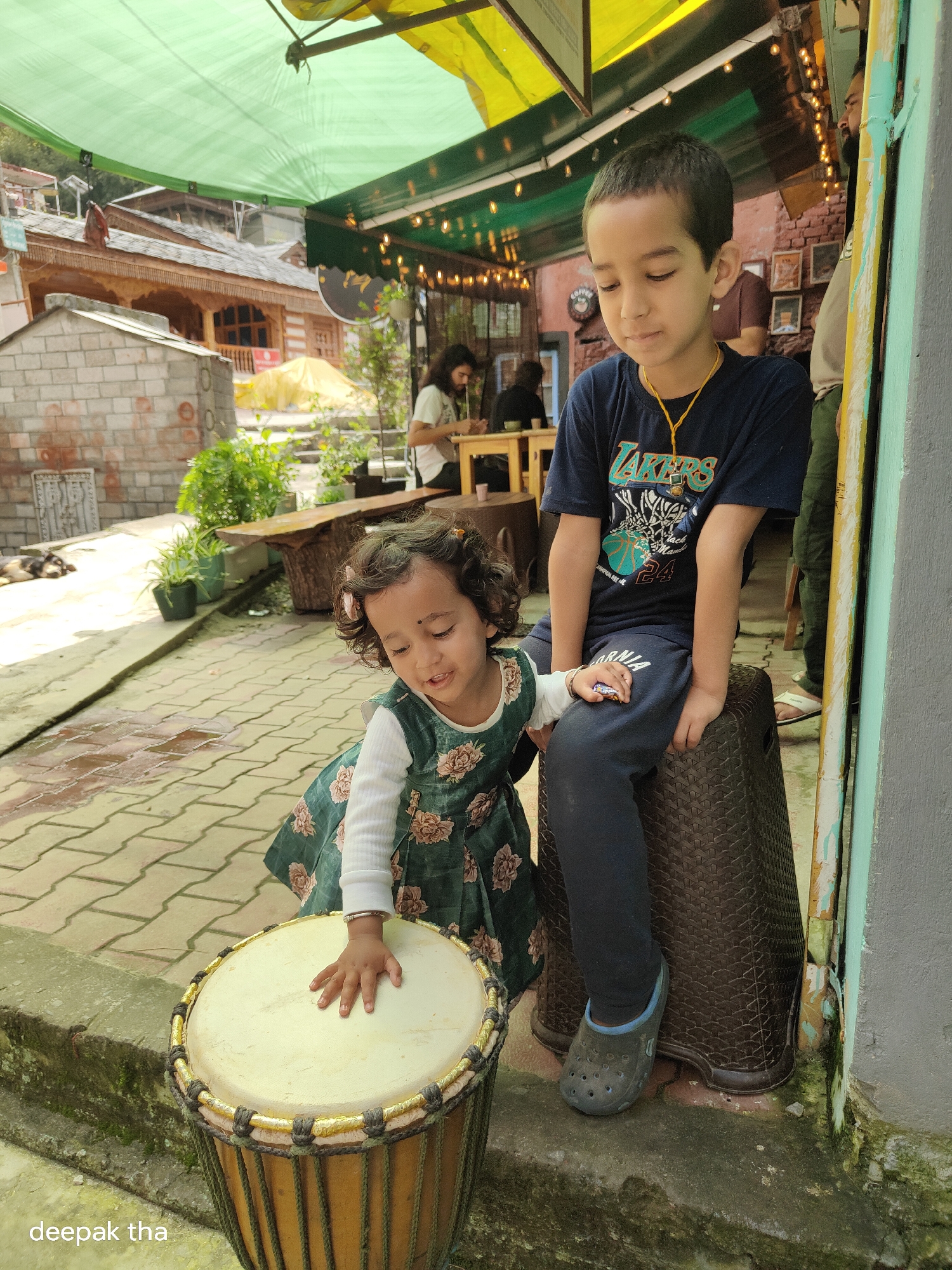 In Vashisht, India, a young boy in a navy blue t-shirt and dark pants is seated on a dark brown, textured plastic stool. He is looking down with a thoughtful expression. A young girl with curly hair, wearing a light-colored long-sleeved shirt and a dark green floral dress, is positioned in front of him. Her right hand is resting on a large, light-colored drum, which has a woven rim with gold and dark green accents. The girl appears to be in the process of playing the drum. They are situated outdoors on a paved area with some wet patches, suggesting recent rain. In the background, there are potted plants, wooden benches, and traditional Indian architecture with a green awning overhead. A dog is visible sleeping on the ground to the left.