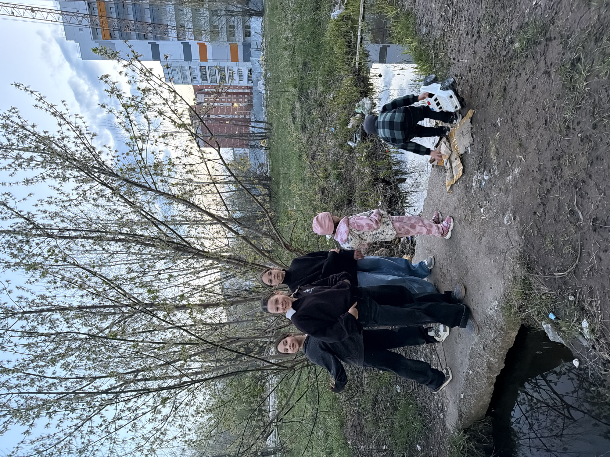 The image shows a group of people in an outdoor setting, likely in or near the city of Chaikovsky, Russia, given the architectural style of the buildings in the background. Four people are standing relatively close together near a bare tree with fresh green leaves, posing for the camera, and two people in the background are involved in an activity along a stream of water.
Three of the people in the foreground appear to be teenagers or young adults; they are all wearing dark clothing, and one is wearing blue jeans. A younger child in a pink hat and patterned jacket stands beside them. In the background, a person in a green and black plaid shirt is crouched down beside a body of water, and may be interacting with a toy and cardboard. The ground is muddy, and there is visible trash near the water's edge.
Behind the group in the foreground, there is a grassy area and a tall building under construction, with a crane. The sky is partly cloudy. The foreground is slightly out of focus.