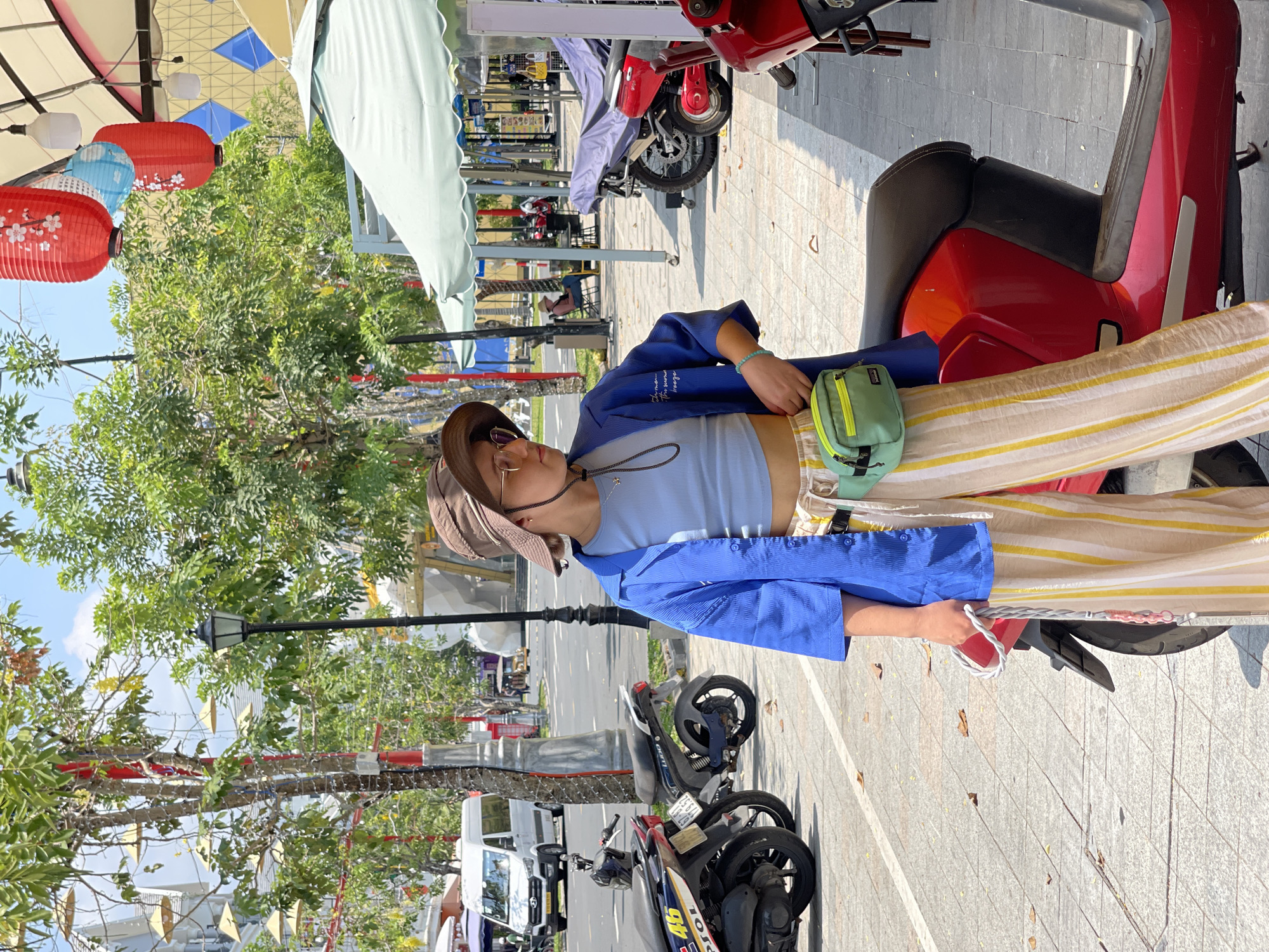 This outdoor image captures a sunny street scene, likely in Phu Quoc, Vietnam. A young person, appearing female, is the central subject, standing or leaning casually beside a bright red scooter. She wears a blue button-up shirt over a light blue ribbed crop top, striped beige and yellow wide-leg pants, and a brown sun hat paired with sunglasses. A light green fanny pack is slung across her chest, and she holds a small white bag with a red cord in her right hand.
The background reveals a bustling street or market area, with multiple scooters and motorcycles parked along the paved road. Overhead, red and light blue paper lanterns, some decorated with white floral motifs, hang from wires beneath a light-colored canopy or awning that features a geometric pattern. Green trees line the street, providing shade, and a black lamppost stands in the mid-ground. The bright sunlight casts distinct shadows, indicating midday or early afternoon. A white van is visible further down the street. Some indistinct white text is visible on the person's blue shirt, and a "96" is seen on a sticker on a motorcycle in the background.