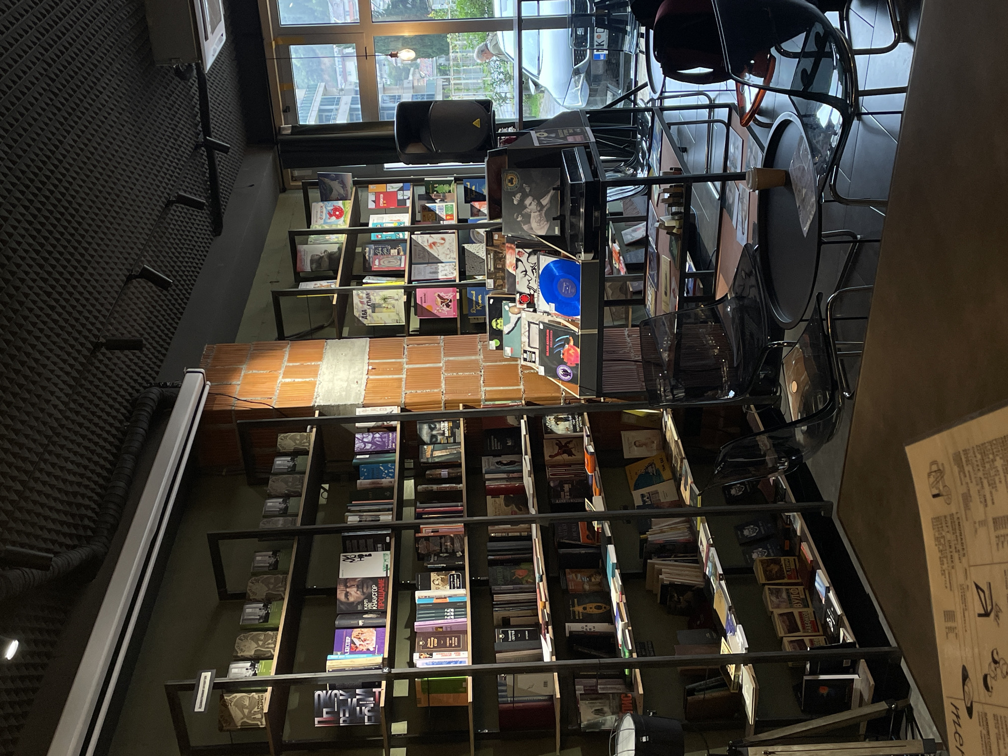 The image shows the interior of a bookstore in Budva, Montenegro. Shelves are filled with books and vinyl records. There are chairs and tables near a window. A car is visible through the window. The bookstore has a modern design with a brick wall.