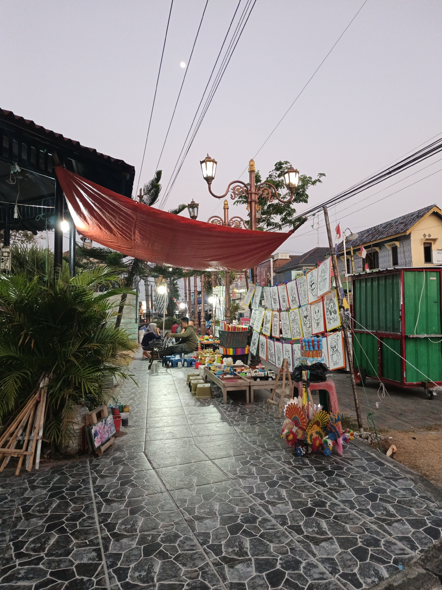 The image depicts an outdoor market scene at dusk in Cawas, Indonesia. The foreground is dominated by a textured, stone-paved walkway. To the left, a large, red tarp is stretched over a makeshift stall, partially obscuring a lush green plant. In the middle ground, two individuals are seated at a small table, one appearing to be an artist sketching or drawing, with several art supplies visible. Several stalls are set up along the walkway displaying various items for sale, including coloring pages featuring cartoon characters, colorful toys resembling birds, and other trinkets. A few small plastic chairs are arranged around some of the stalls. In the background, a yellow building with a dark roof is visible, along with a tall, green shipping container on wheels. Decorative streetlights with ornate metalwork are positioned along the pathway, and overhead, a network of power lines stretches across the pale sky. The moon is faintly visible in the upper left quadrant of the image. The overall atmosphere suggests a quiet evening at a local marketplace.