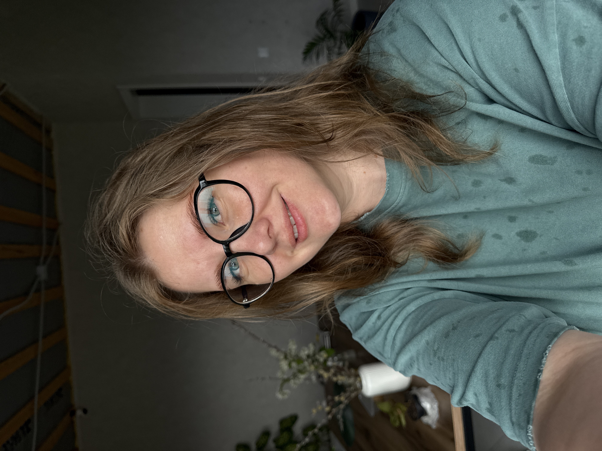 A woman with light brown hair and blue eyes is wearing round black-rimmed glasses and a light green, long-sleeved shirt. She is looking directly at the camera and smiling slightly. The location is Ферма, Russia. The background is a dimly lit room with a light fixture near the ceiling and a plant in the upper right corner. In the lower part of the image, there are more plants and some blurred objects.