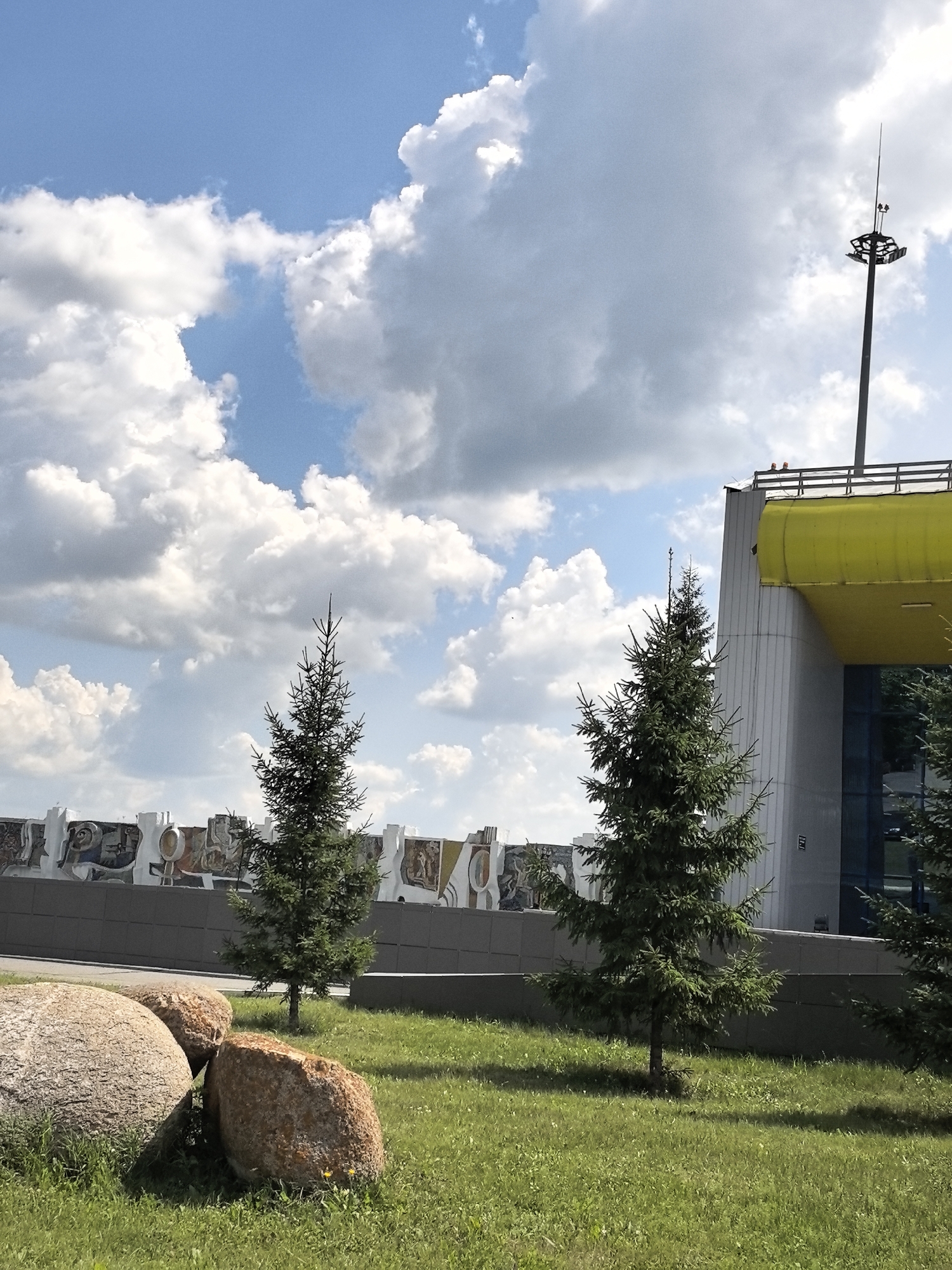 This is a landscape photograph of an outdoor scene on a sunny day. In the foreground, on a grassy area, are three large, round, textured rocks. Behind the rocks, a few young coniferous trees are visible, with a larger one to the right of the frame. A long, low wall with a mosaic or mural decoration runs across the mid-ground, separating the grassy area from a modern building in the background. This building has a prominent yellow-colored overhang and a grey, angular facade. A tall pole with lights and what appears to be a weather station or antenna stands near the top right corner of the image, behind the building. The sky is blue with scattered white, fluffy clouds. The image appears to be taken in the Прибрежный ауылдық округі, Kazakhstan, possibly at an airport or a similar public facility, given the lighting tower and modern architecture. No people are visible in the image.
