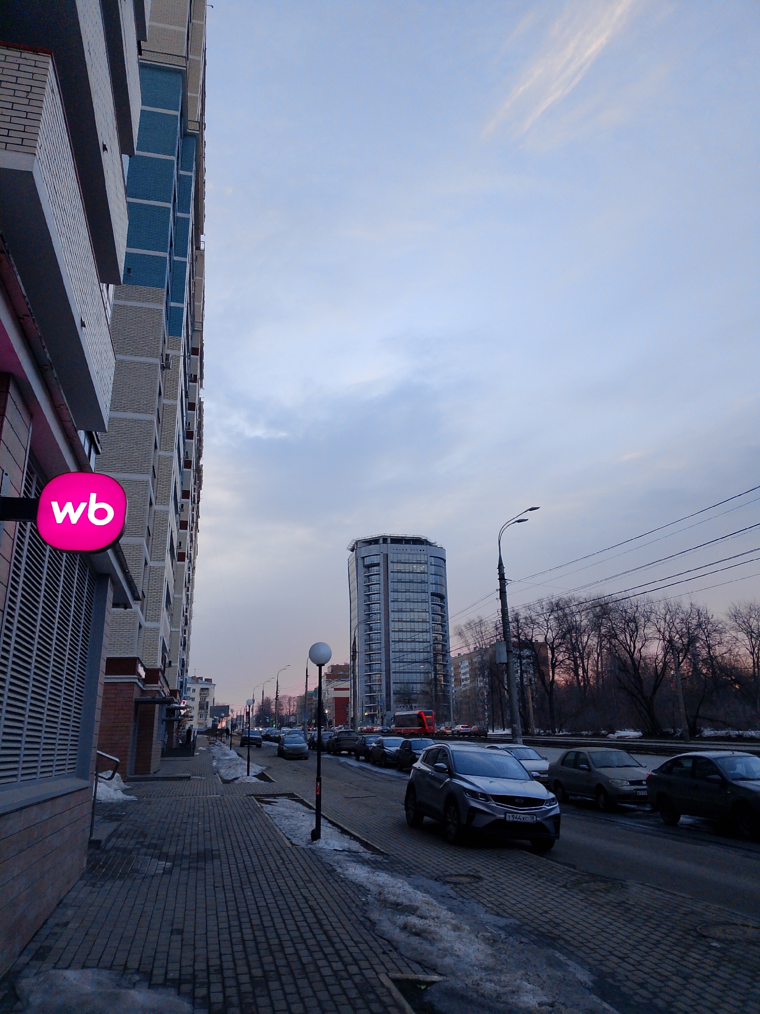 The image depicts a street scene in Izhevsk, Russia, with a view down a city street. In the foreground, a tall building with a pink "WB" (likely Wildberries) sign is on the left. A paved sidewalk runs alongside the building.
The street has multiple lanes of traffic with several parked and moving cars. A prominent modern skyscraper stands in the middle distance. A row of bare trees lines the street on the right, and beyond them, the sky is overcast with hints of pink. Snow and ice patches are visible along the curb, indicating a cold climate. Streetlights are evenly spaced along the road.