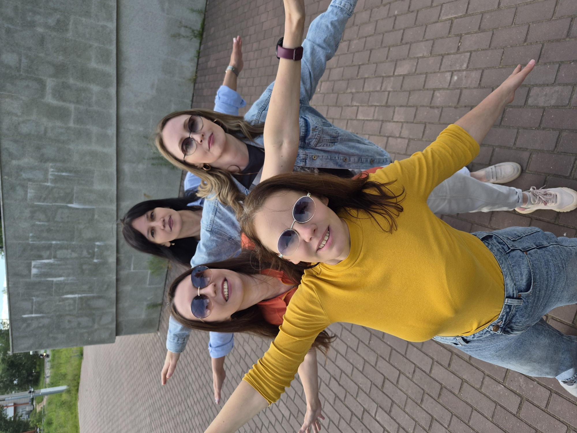 The image features four adult women posing outdoors with a tilted camera angle, making them appear to be leaning or suspended with their arms outstretched horizontally. They are arranged diagonally across the frame, from the bottom-right to the top-left, and all are smiling towards the camera.
The woman closest to the foreground, at the bottom-right, has long brown hair and is wearing a yellow long-sleeved top, blue jeans, and light-colored sneakers. She sports round, mirrored sunglasses. Next to her, towards the center, is a woman with long dark brown hair, an orange top, blue jeans, and similar light-colored sneakers, also wearing round, mirrored sunglasses.
Further back, a woman with blonde or light brown hair wears a light blue denim jacket over a dark top, blue jeans, and light sneakers. She has aviator-style sunglasses and a dark-colored smartwatch on her left wrist. The fourth woman, at the top-left, has dark hair and is dressed in a light blue denim jacket over a dark top, light-colored pants, and light sneakers, without visible sunglasses.
The ground beneath them is paved with reddish-brown bricks. In the background, there is a large, grey concrete wall or structure, suggesting an urban or park environment. Some sparse greenery is visible behind the concrete structure, along with hints of distant elements like roads or buildings in the far background.
The setting appears to be daytime, with bright, even lighting, implying clear weather. All the women display cheerful and relaxed expressions, indicating a positive mood. There is no visible text in the image.