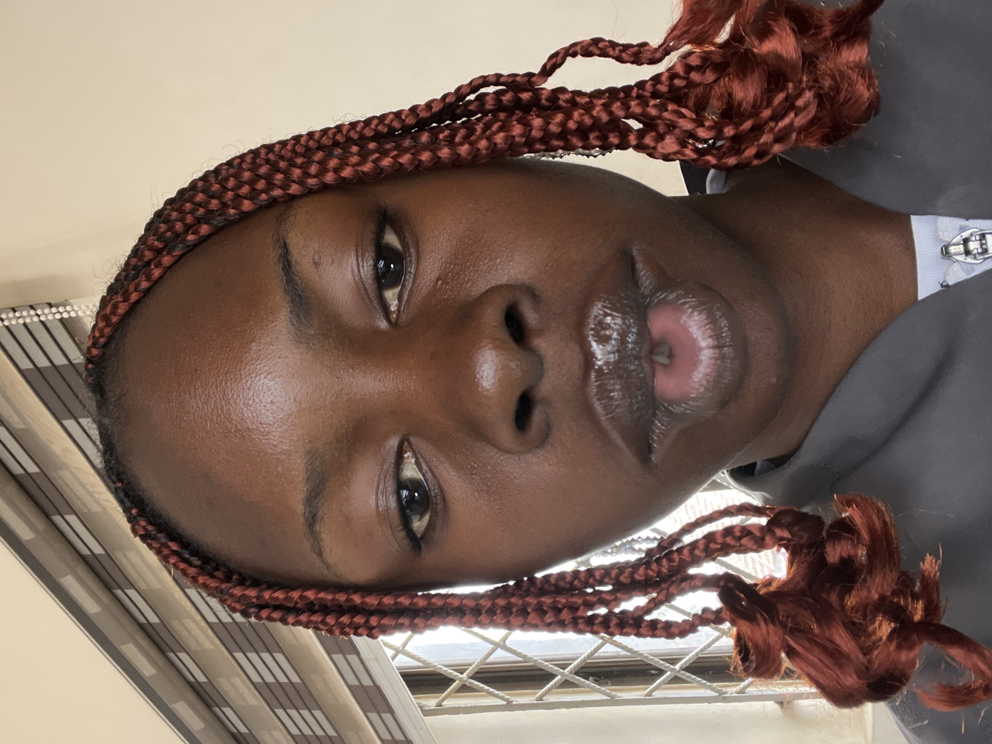 The image is a close-up selfie of a young Black woman with dark skin and braided hair. The braids are a reddish-brown color and are styled in cornrows that extend down the sides of her face. Her eyes are brown, and she is wearing lip gloss. She is wearing a gray jacket over a white shirt. The background appears to be indoors, with blinds visible behind her. The overall lighting suggests it is daytime. The image does not contain any information about the city of Jos, Nigeria, nor does it depict any specific city landmarks or environmental features that would allow for its localization.