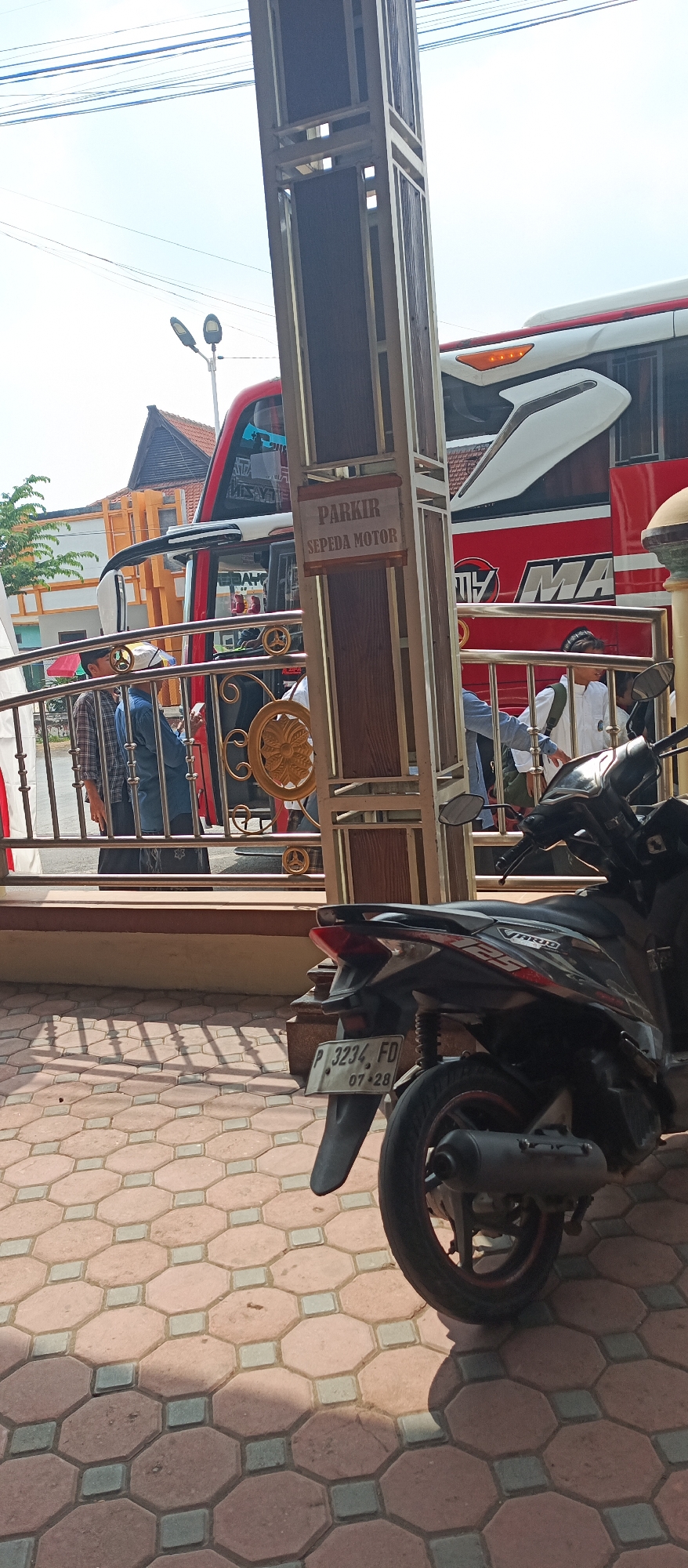 In the city of Kapongan, Indonesia, a motorcycle is parked on a patterned pavement. In the background, a red and white bus is visible, with a sign on a pillar that reads "PARKIR SEPEDA MOTOR" (motorcycle parking). Several people are near the bus and a metal railing. The scene is outdoors, with power lines and a street lamp visible in the sky. The motorcycle's license plate reads "P 3234 FD 07-28".
