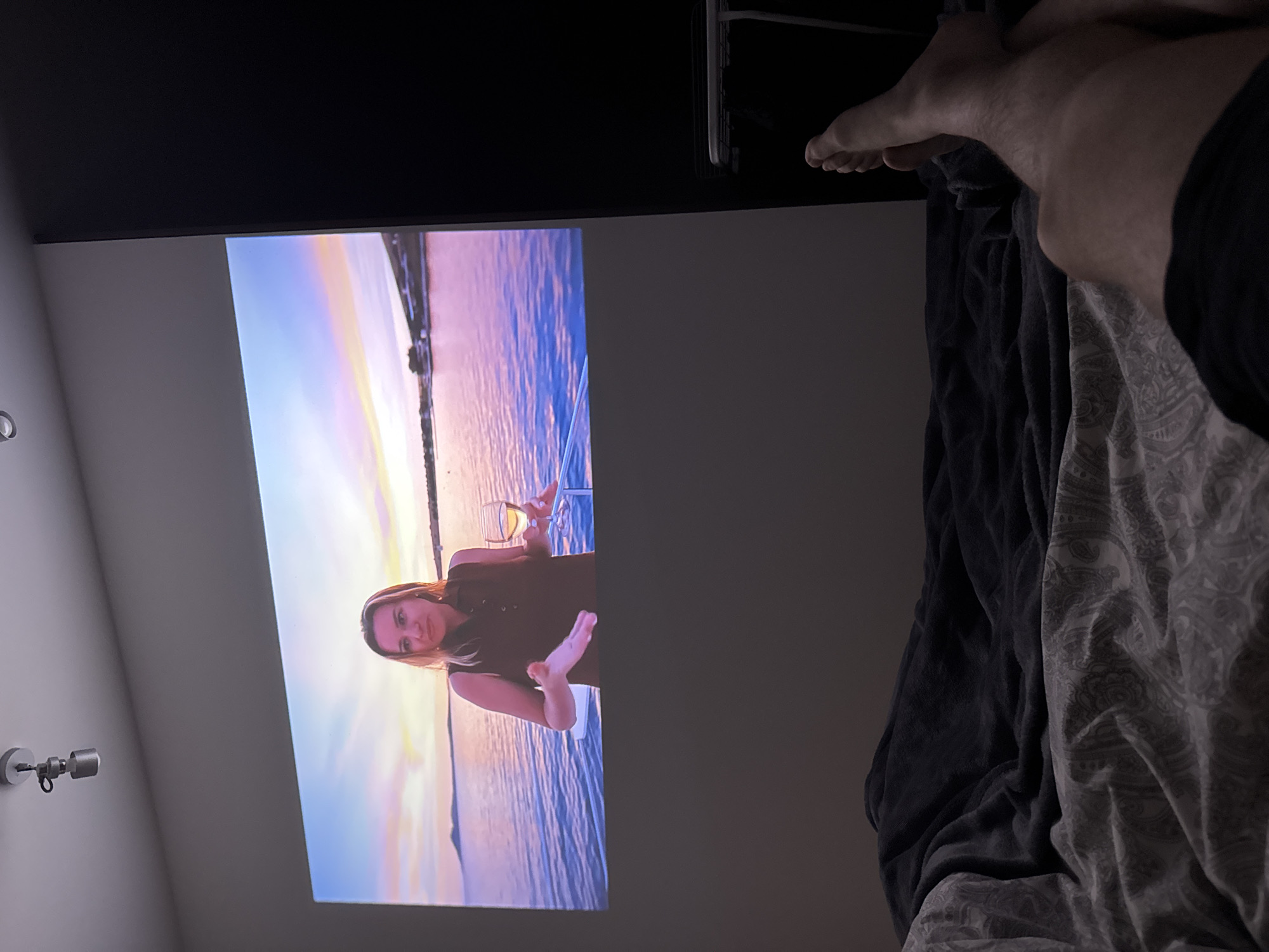 The image shows a partially visible human lying on a bed, with feet and legs exposed. The bedding consists of patterned sheets and a dark blanket. Above the bed, a projector displays an image of a woman holding a wine glass, set against a sunset-like backdrop. The projector's image is displayed on what appears to be a light-colored wall. On the left side of the wall, two cylindrical light fixtures are mounted. A black cabinet or shelf is partially visible above the projected image.
There is no information that indicates the location is in Warsaw, Poland.