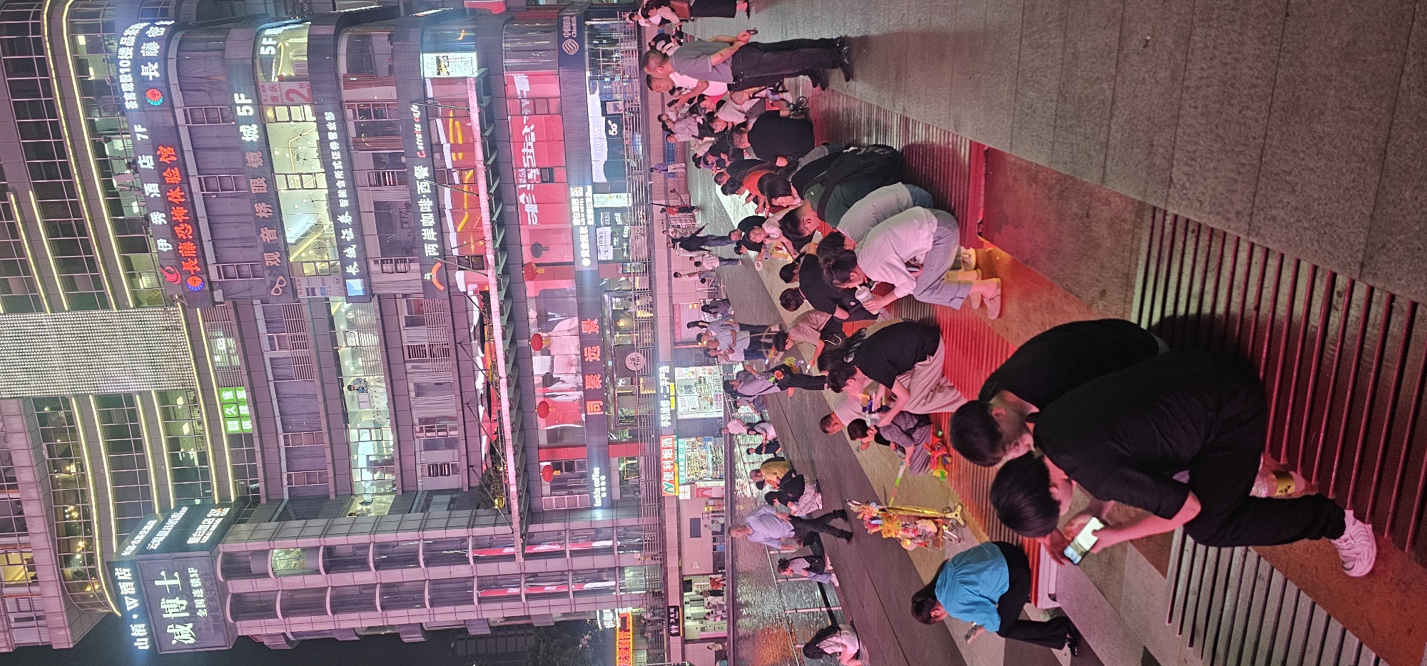 In Liangjiang, China, a group of people are kneeling on a walkway at night. Most of them are using their phones. Behind them are tall buildings with signs, lights, and text in Chinese. The architecture includes repeating rectangular windows and rounded edges.