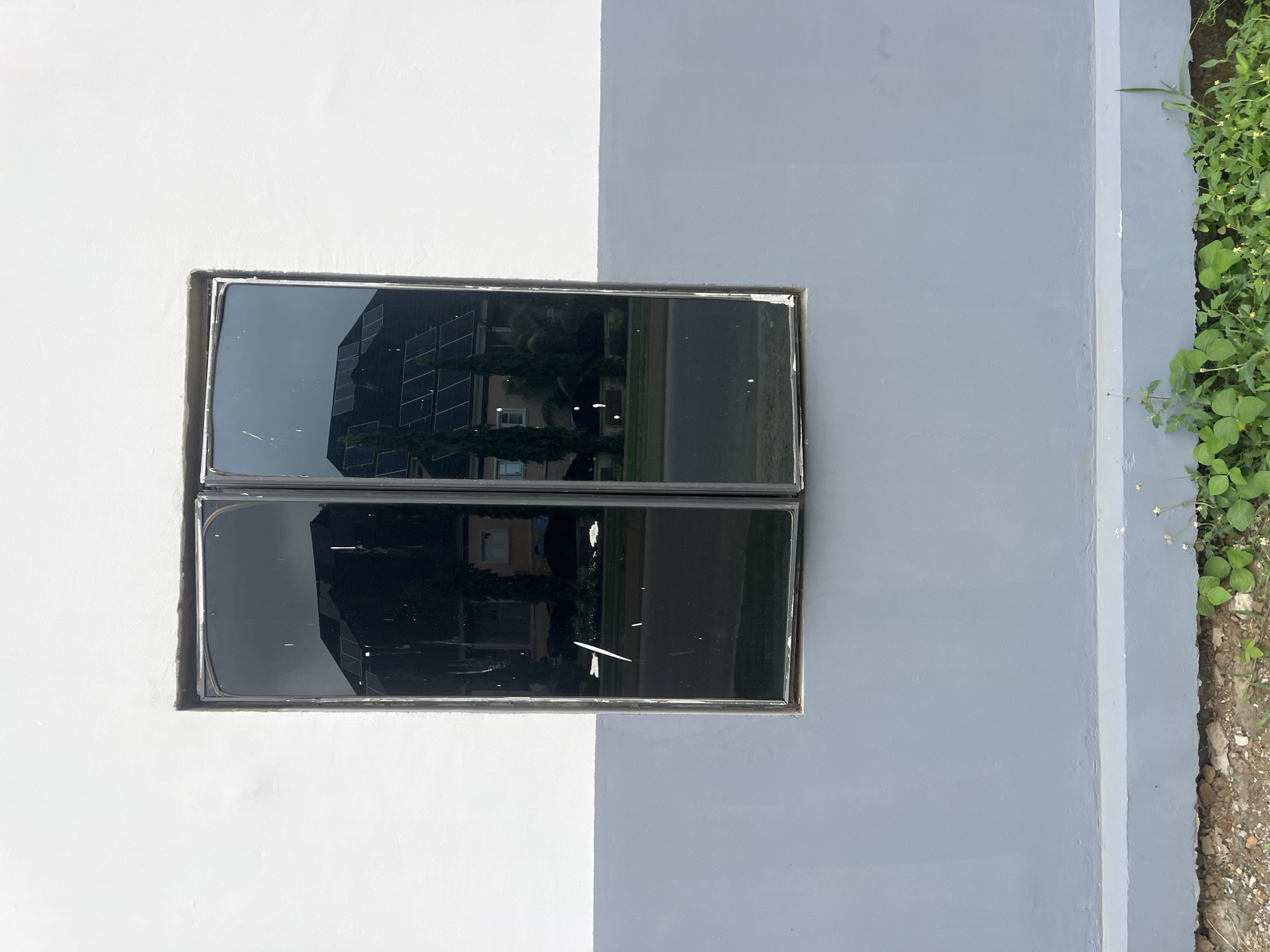 This media file displays a close-up view of a wall painted in two distinct colors: white on the left and a lighter shade of gray on the right. Embedded within the wall is a rectangular window with dark, reflective glass panes. The window is divided horizontally, with two panes stacked vertically, creating a grid-like appearance. The reflections in the glass reveal a modern building with multiple floors and a facade that appears to be composed of glass panels and possibly solar panels. Green foliage, likely trees or bushes, is also visible in the reflection, suggesting an outdoor environment.
To the right of the gray section of the wall, a small patch of green vegetation grows alongside the wall, with small white flowers interspersed among the leaves. The ground beneath the vegetation is composed of dirt and small stones. The overall scene suggests an exterior wall of a building, possibly in an urban or suburban setting, given the reflection of the multi-story building. The lighting in the image indicates daylight, likely midday, with the sun casting a bright illumination on the wall. There is no discernible text in the image. The location context is identified as Karum, Nigeria.