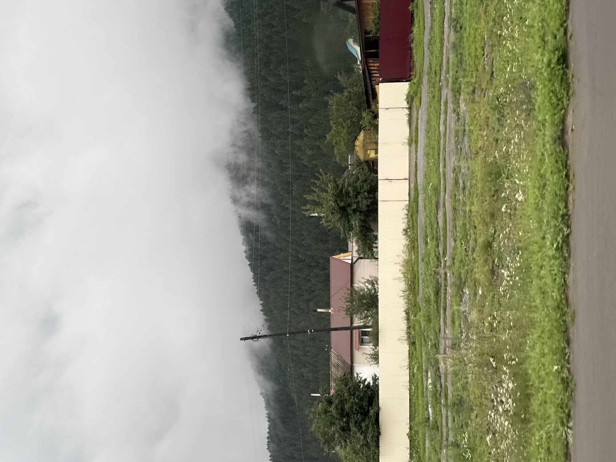 The image depicts a rural scene in Elekmonar, Russia, characterized by a overcast sky and a forested hillside partially shrouded in mist. In the foreground, a beige fence runs horizontally across the frame, separating a grassy area with wildflowers from what appears to be a dirt or gravel road. Beyond the fence, several buildings are visible. One structure has a light-colored exterior with a prominent red roof and a single visible window. A utility pole stands near this building. Further to the right, a wooden structure with a dark red fence is partially visible, along with a smaller yellow greenhouse-like structure. The dense green forest dominates the background, climbing the hillside and disappearing into the clouds and fog. The overall atmosphere is subdued and tranquil, suggesting a cool or damp day.