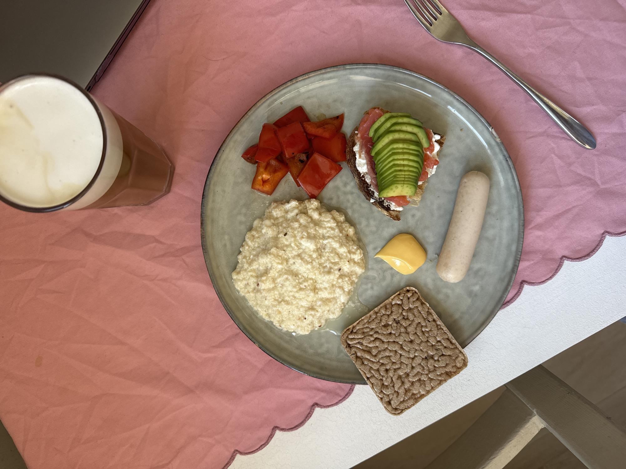 A top-down shot depicts a breakfast plate arranged on a white table. The plate holds: diced red peppers, a mound of cooked grains (perhaps oatmeal), a dollop of yellow sauce, a sausage, a textured, square-shaped crispbread, and a slice of bread topped with cream cheese, salmon, and avocado. A silver fork lies to the right of the plate. A glass of frothy beverage is positioned to the left, partially obscured by a laptop. A light pink tablecloth provides a backdrop. The scene is brightly lit. The location cannot be determined with the available information.