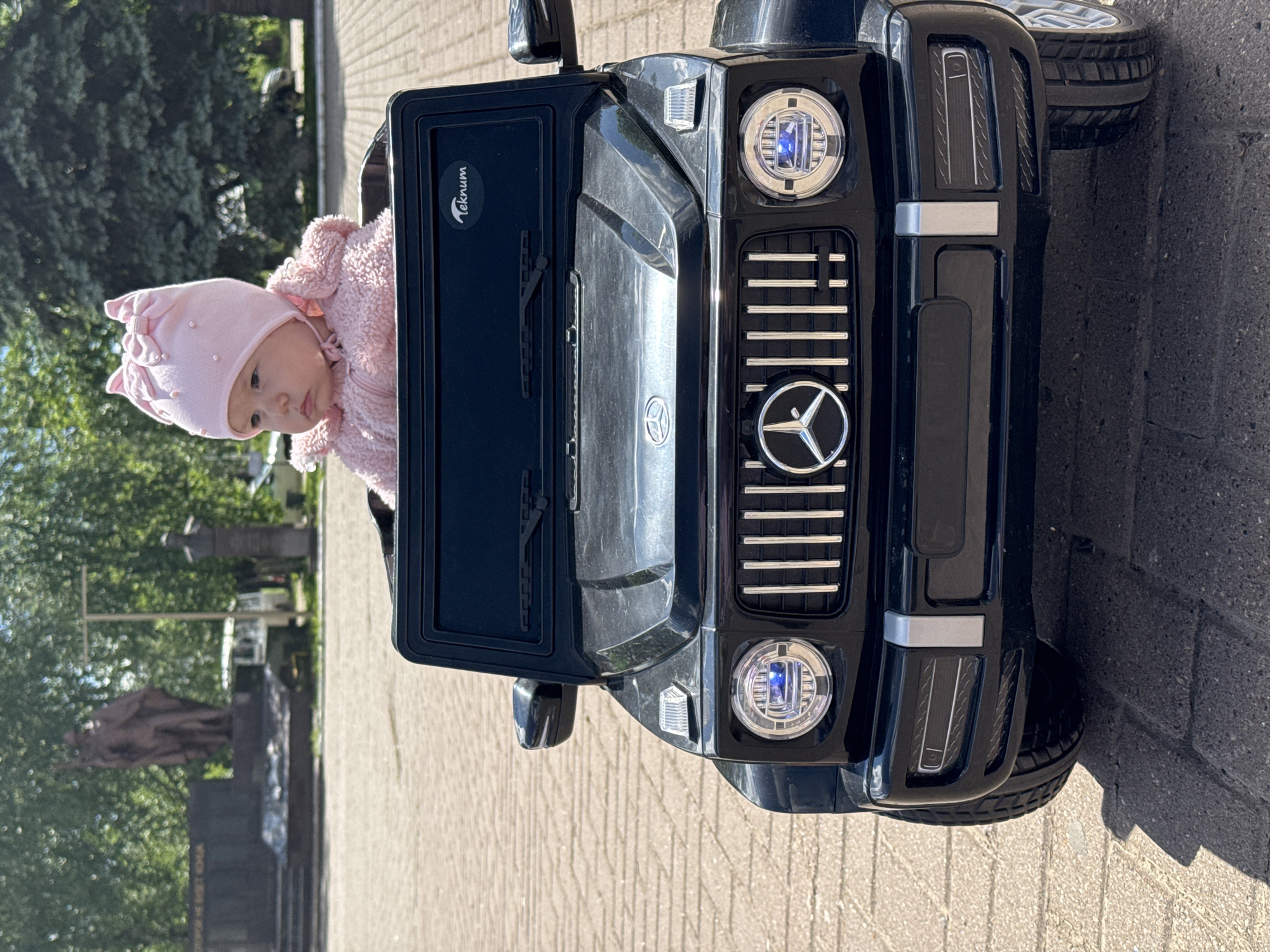 A baby wearing a pink hat and pink sweater is in a black Mercedes-Benz toy car. The location is Zav'yalovo, Russia, judging by the monument visible in the background. The car is on a brick-paved surface.