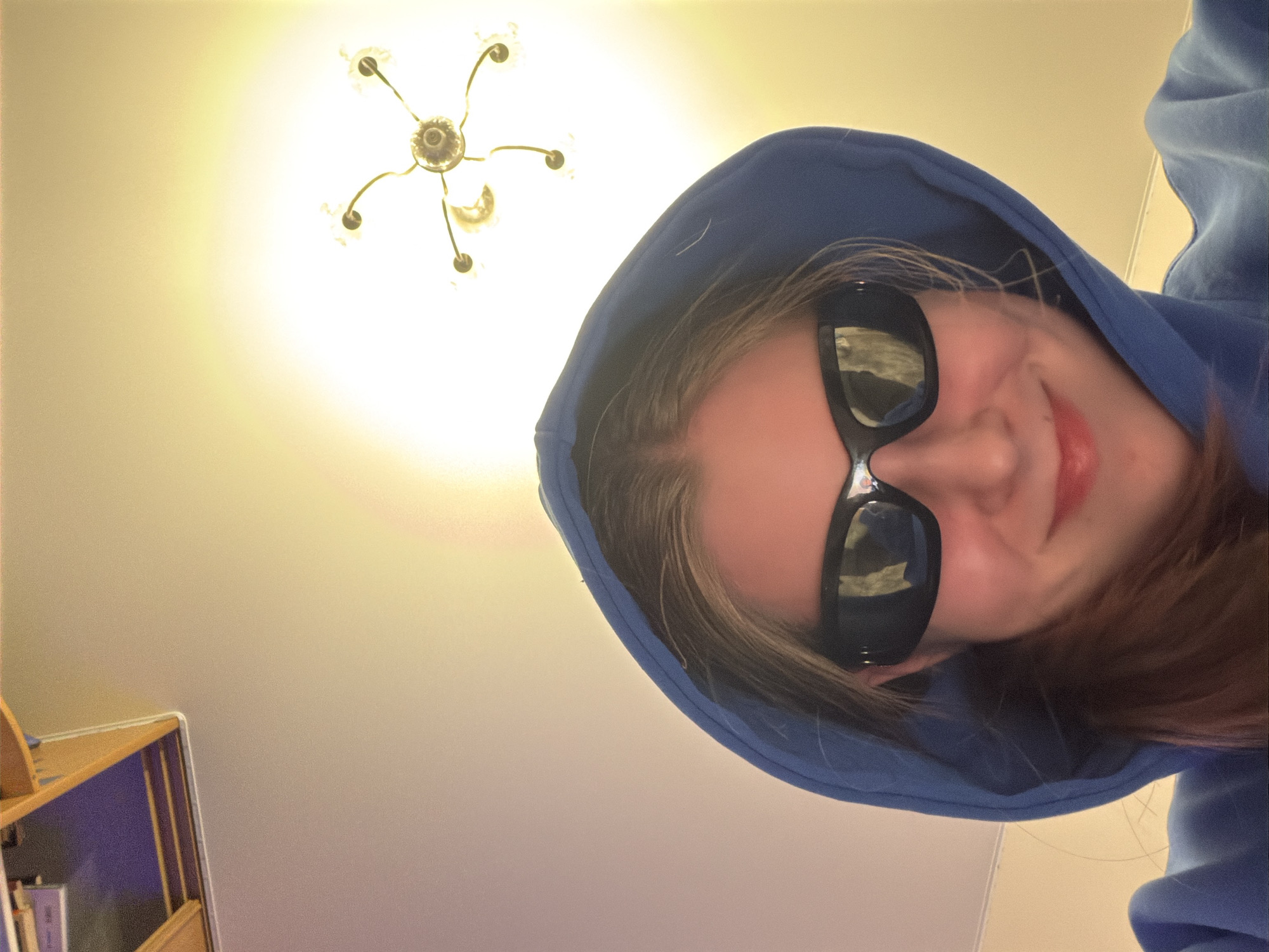 This close-up image, taken in Perm, Russia, features a person looking upwards towards the camera. The individual is wearing a blue hooded sweatshirt, with the hood pulled over their head, and dark, rectangular sunglasses that reflect a distorted view of the surroundings. A subtle smile is visible on the person's mouth, indicating a cheerful or playful expression. Their hair, light brown or blonde, is visible around their face and beneath the hood.
The scene is set indoors. In the upper left background, a multi-armed chandelier with illuminated bulbs emits a bright, warm light, dominating the ceiling area. The ceiling and upper walls appear to be a light, warm beige color. A small portion of a wooden shelf or cabinet is visible in the lower left corner, suggesting a furnished room. The primary light source is artificial, originating from the chandelier, creating a bright but warm ambiance. The overall mood is casual and lighthearted. No discernible text is visible in the image.