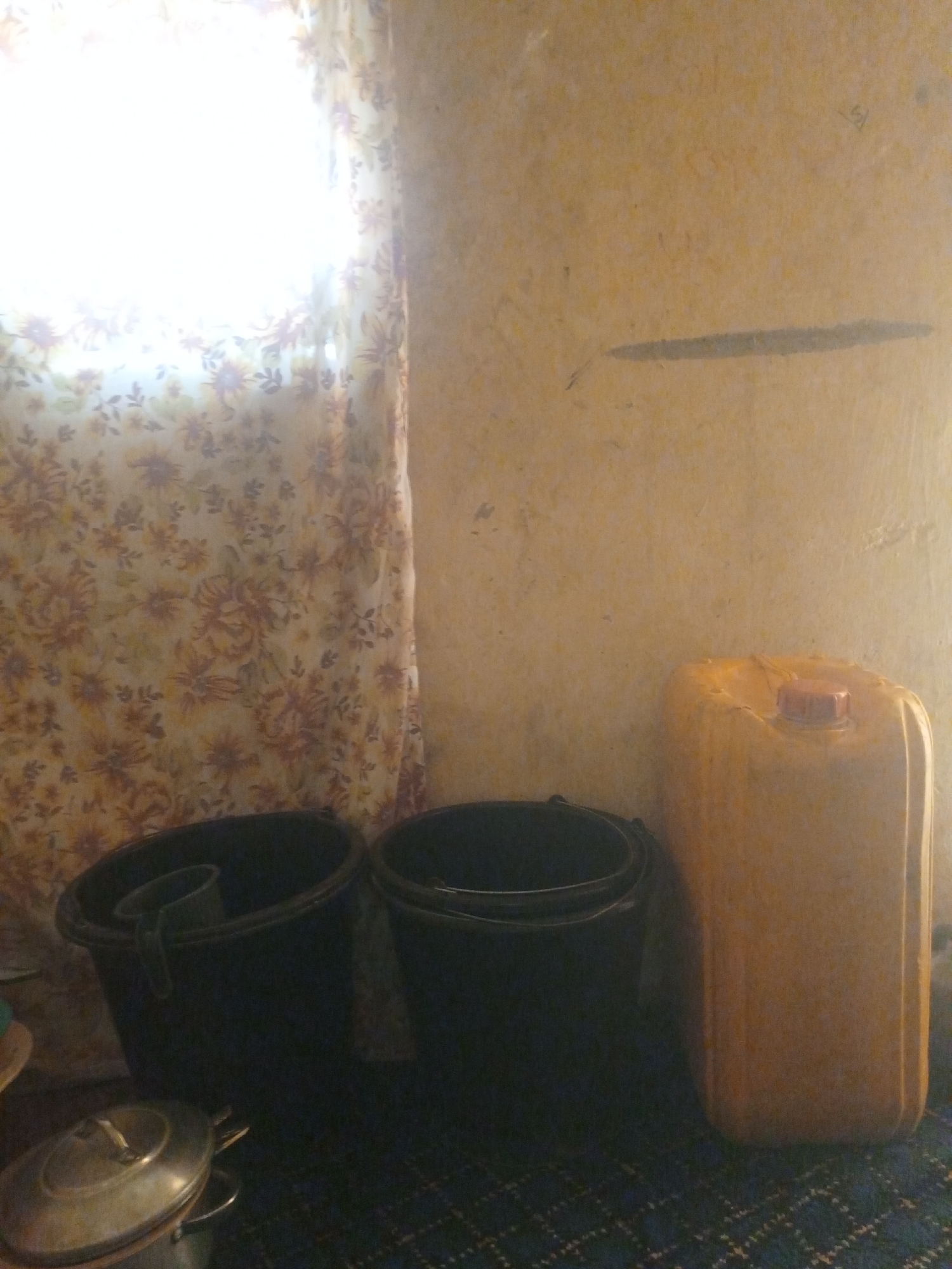 In Jalingo, Nigeria, an interior scene depicts a corner of a room with a window covered by a floral patterned curtain, allowing bright light to filter through. To the right of the curtain stands a large, translucent yellow plastic jerrycan with a red cap. In front of the jerrycan and to the left, two dark-colored buckets are visible. One bucket, positioned further left, contains a smaller, lighter-colored cup. In the foreground, partially visible, are metallic pots, one with a lid. The floor is covered with a patterned rug. The wall is a muted yellow color with a faint horizontal mark.