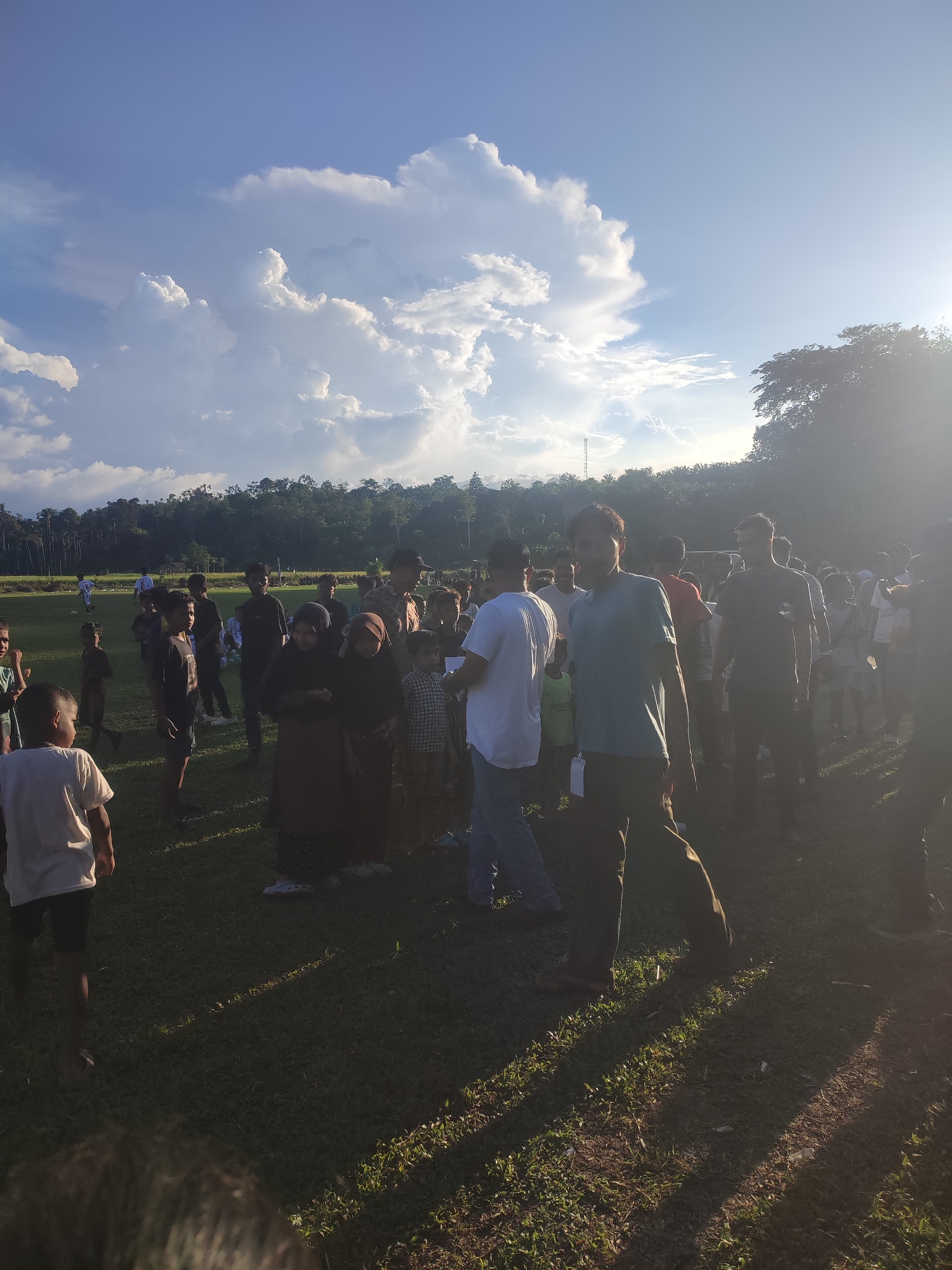 A group of people, including children, are gathered on a grassy field under a partly cloudy sky. The background shows trees. The location is Babahlueng, Indonesia. People are standing and interacting. The sun is shining, casting long shadows on the grass. Some individuals are wearing traditional Muslim clothing.