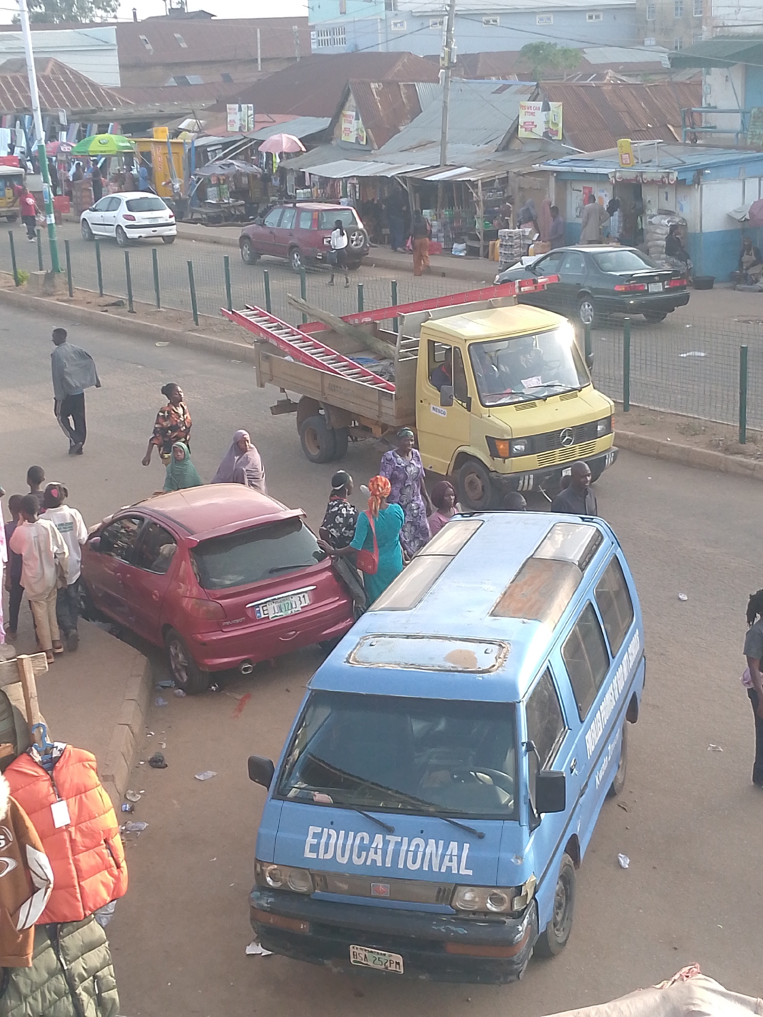 This is an aerial, slightly angled, medium shot of a street scene in Bukuru, Nigeria, likely taken during the day under natural light. The image captures a busy urban environment with several vehicles and pedestrians.
In the foreground, a blue minibus with "EDUCATIONAL" written on its windshield is parked on the right side of the street. The minibus appears to be well-used, with some visible wear and tear. To the left of the minibus, a red hatchback car is parked, facing away from the viewer. A group of people are gathered around the red car, with some individuals standing near the rear and others in the vicinity.
Further back, a yellow Mercedes-Benz truck is parked in the street, with a red ladder secured on its flatbed. Several people are also visible around the truck, some appearing to be interacting. Across the street, a row of market stalls with corrugated metal roofs line the road, with awnings and open fronts displaying various goods. Several individuals are seen walking and browsing in front of these stalls. In the background, more buildings with similar tin roofs are visible, suggesting a dense urban settlement. A dark green sedan is driving away from the viewer on the far side of the street, passing by a white hatchback car. The road surface is paved, and a metal fence runs along the side of the street. The overall impression is one of a bustling marketplace and transportation hub.