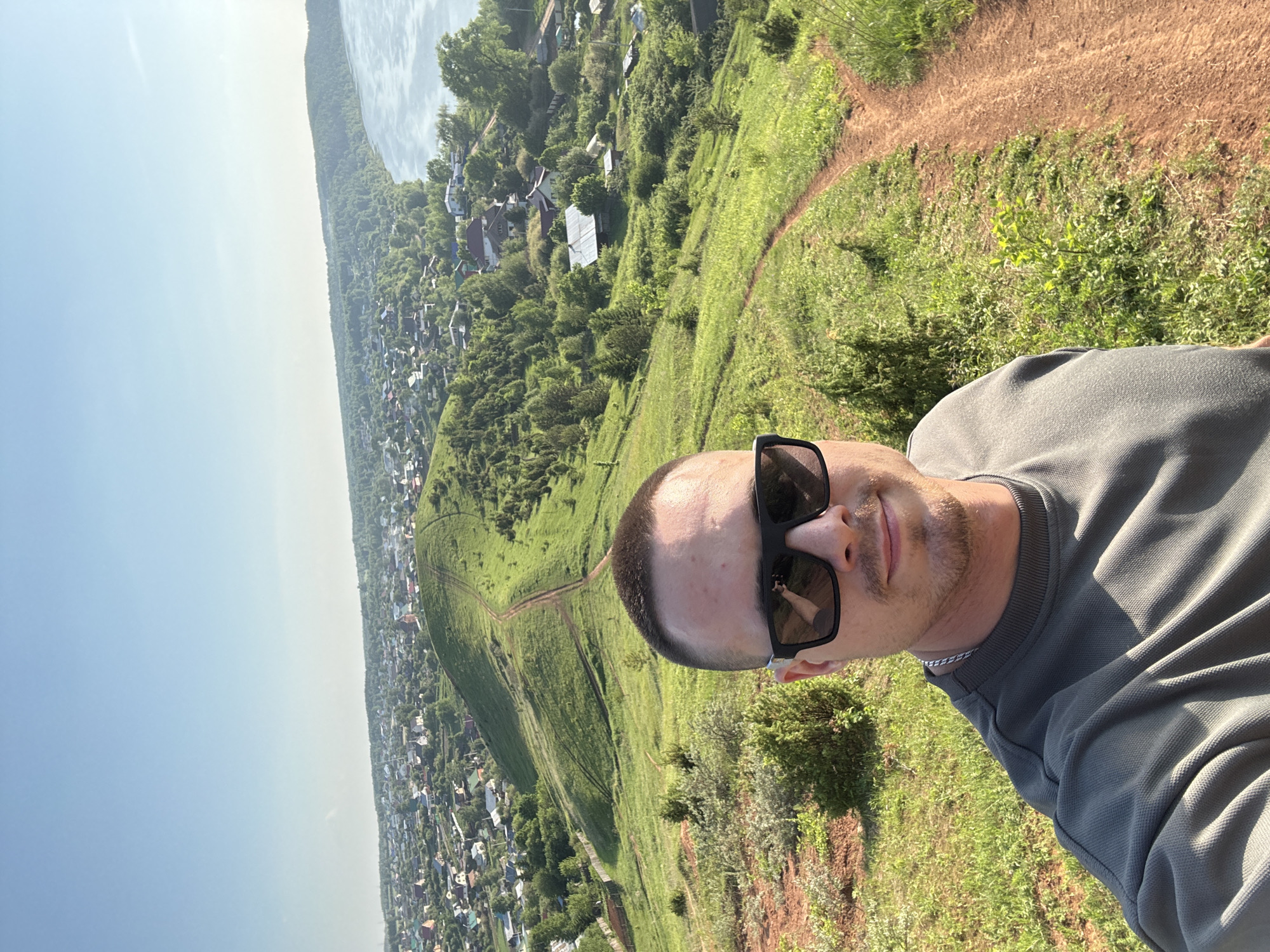 A selfie taken outdoors features a man with a closely cropped haircut and sunglasses. He is wearing a grey collared shirt and a silver chain. The background displays a steep, grassy hill. At the bottom of the hill, the city of Ольховка, Russia, stretches into the distance with numerous buildings and trees. A river borders the town. The sky is clear and bright. A dirt path winds up the hill.