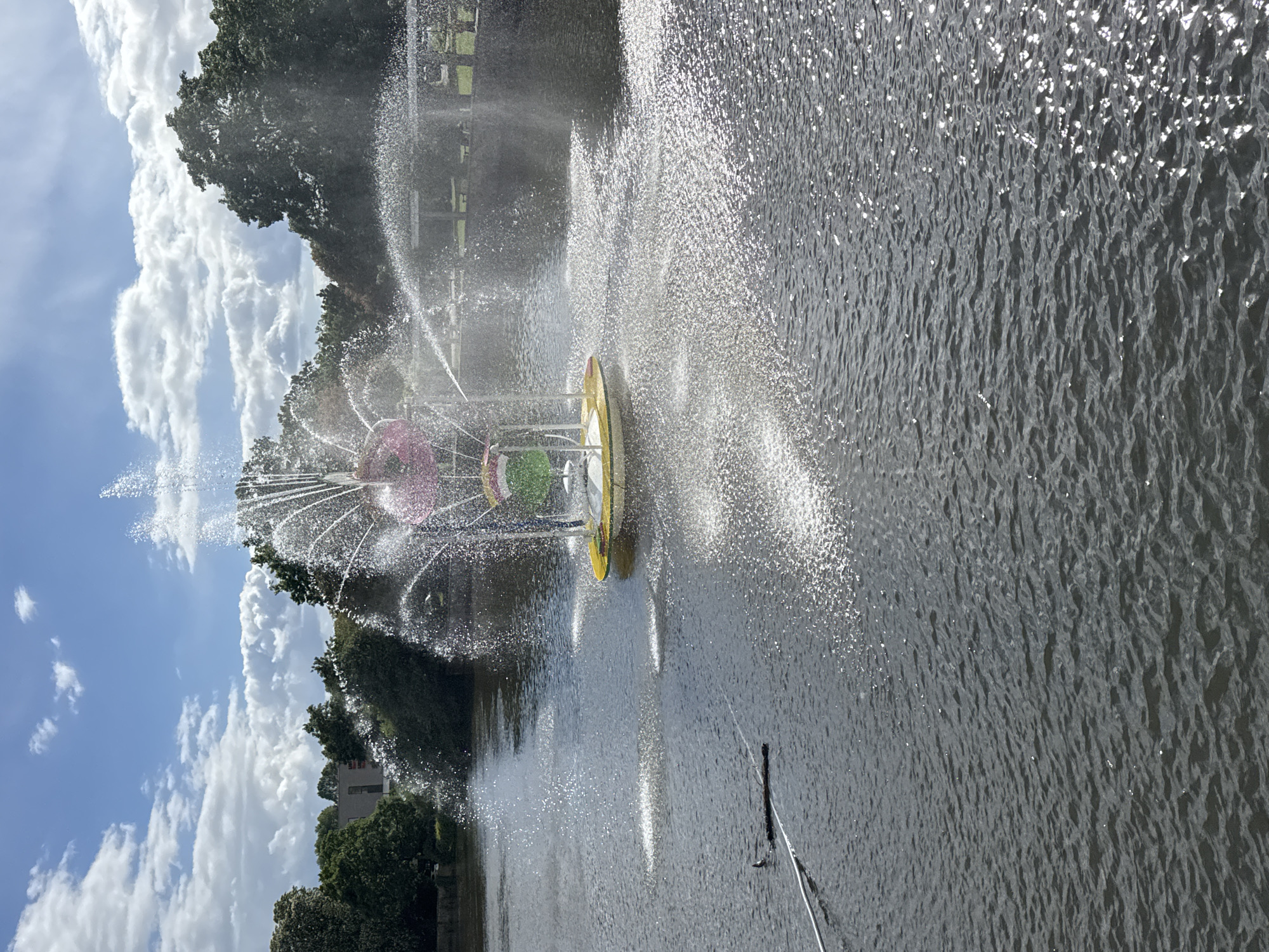The image captures a scene in Bietigheim-Bissingen, Germany, featuring a water fountain within a body of water, likely a pond or lake. The fountain has a wheel structure, with sections painted pink, green, and yellow. Water sprays outwards from the fountain, creating a dynamic, sparkling effect, particularly visible on the right side of the frame where the sunlight reflects off the water droplets. The body of water exhibits ripples and reflections. The surrounding environment includes lush green trees, a partly cloudy sky, and a building partially visible among the trees. The presence of a pole or wire extending from the bottom right suggests the presence of equipment for the fountain.