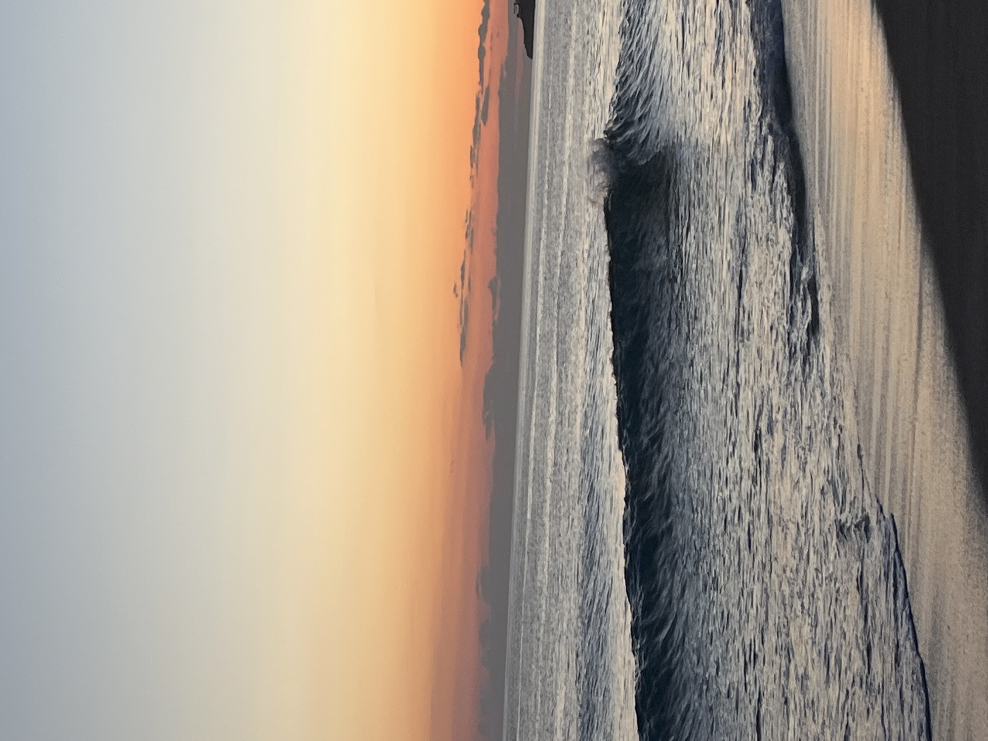 The image captures a seascape at sunset in Alanya, Turkey. The scene presents a wide beach to the right, with a textured pattern indicative of wet sand. Waves are visible along the shore, with water ranging from a dark hue to lighter shades. The sky transitions from a soft orange glow near the horizon to a pale blue above, suggesting dusk or dawn. A thin strip of land or distant clouds are visible near the horizon. The lighting emphasizes a serene and tranquil atmosphere.