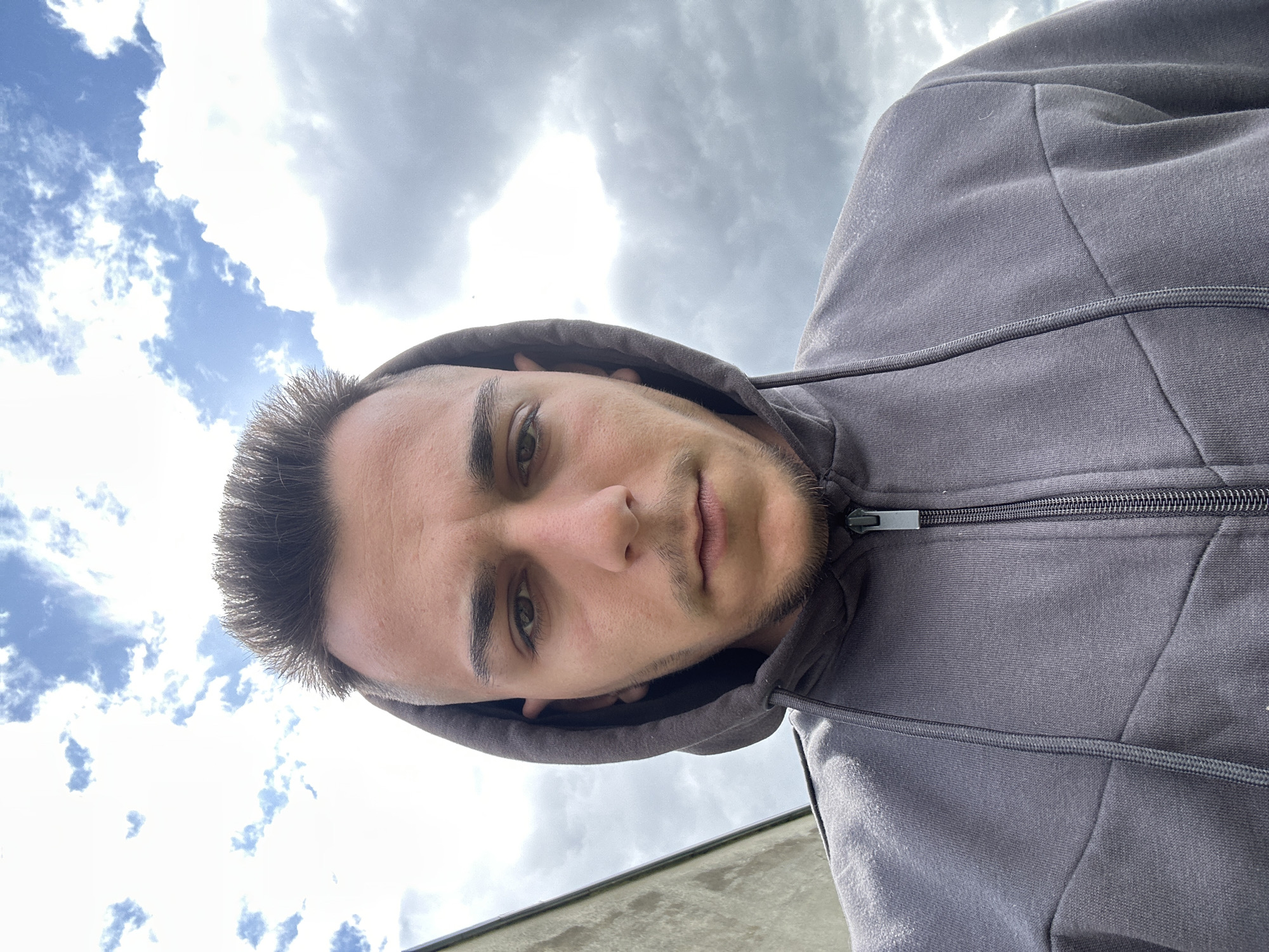 A low-angle, close-up selfie features a male individual wearing a gray hooded sweatshirt with the hood up. The zipper of the sweatshirt is partially visible. The man has short, dark hair, brown eyes, and light facial hair. The background consists of a cloudy sky with patches of blue. The location is Srednia, Bulgaria. There is a light-colored building in the very lower portion of the picture.