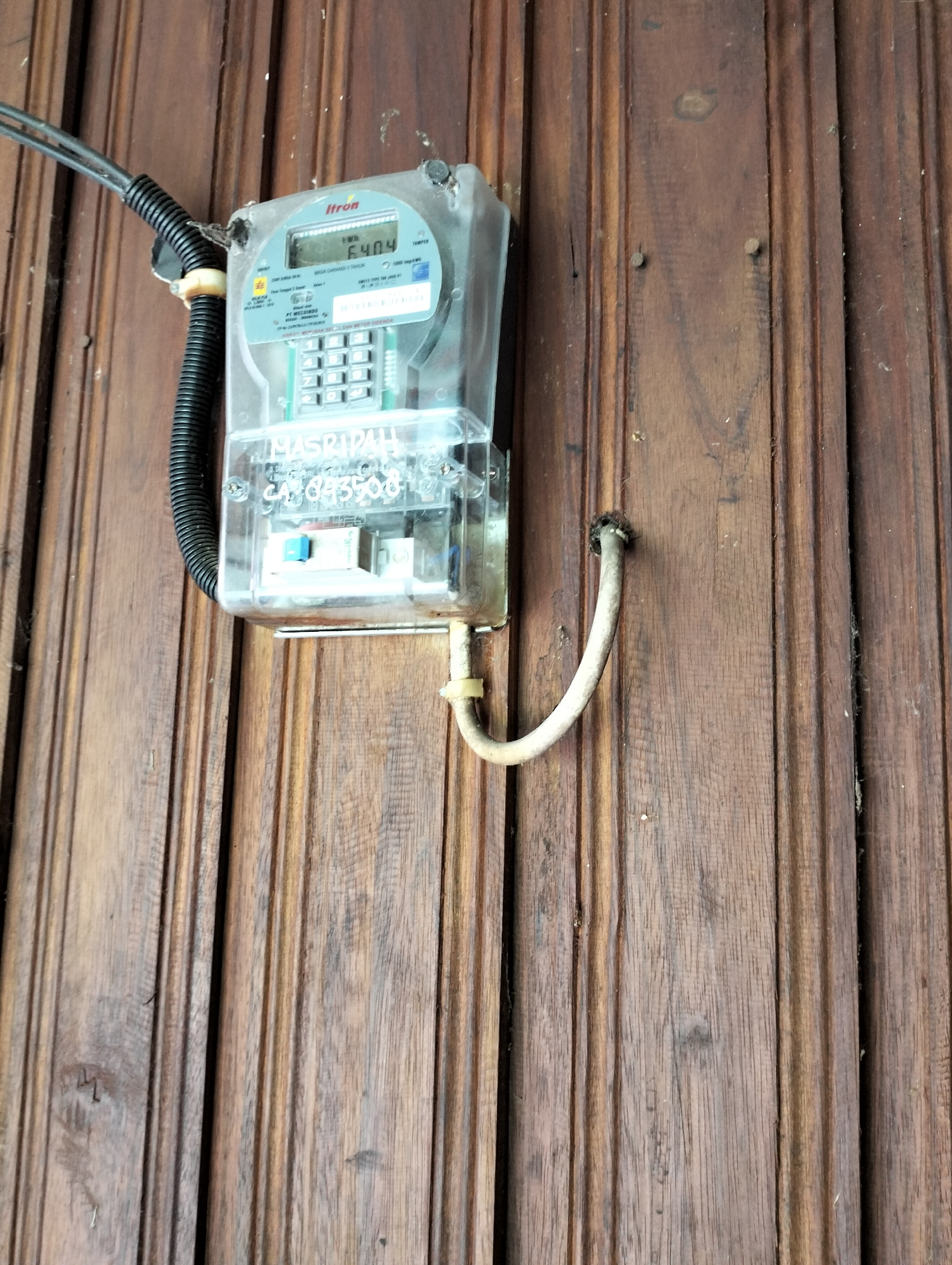 An outdoor shot captures a close-up of an electricity meter mounted on a wooden wall. The meter, encased in clear plastic, displays a digital reading of 6404. The meter is connected to black electrical conduits that enter from the left, and a bent metal pipe extends from the bottom right of the meter and disappears into a hole in the wall. The wooden wall behind the meter features vertical planks with visible grain and a weathered appearance. The background is largely obscured by the meter and the wall. The scene is indicative of an external residential or commercial building setup for electricity supply. The location is identified as Mandala Jaya, Indonesia.