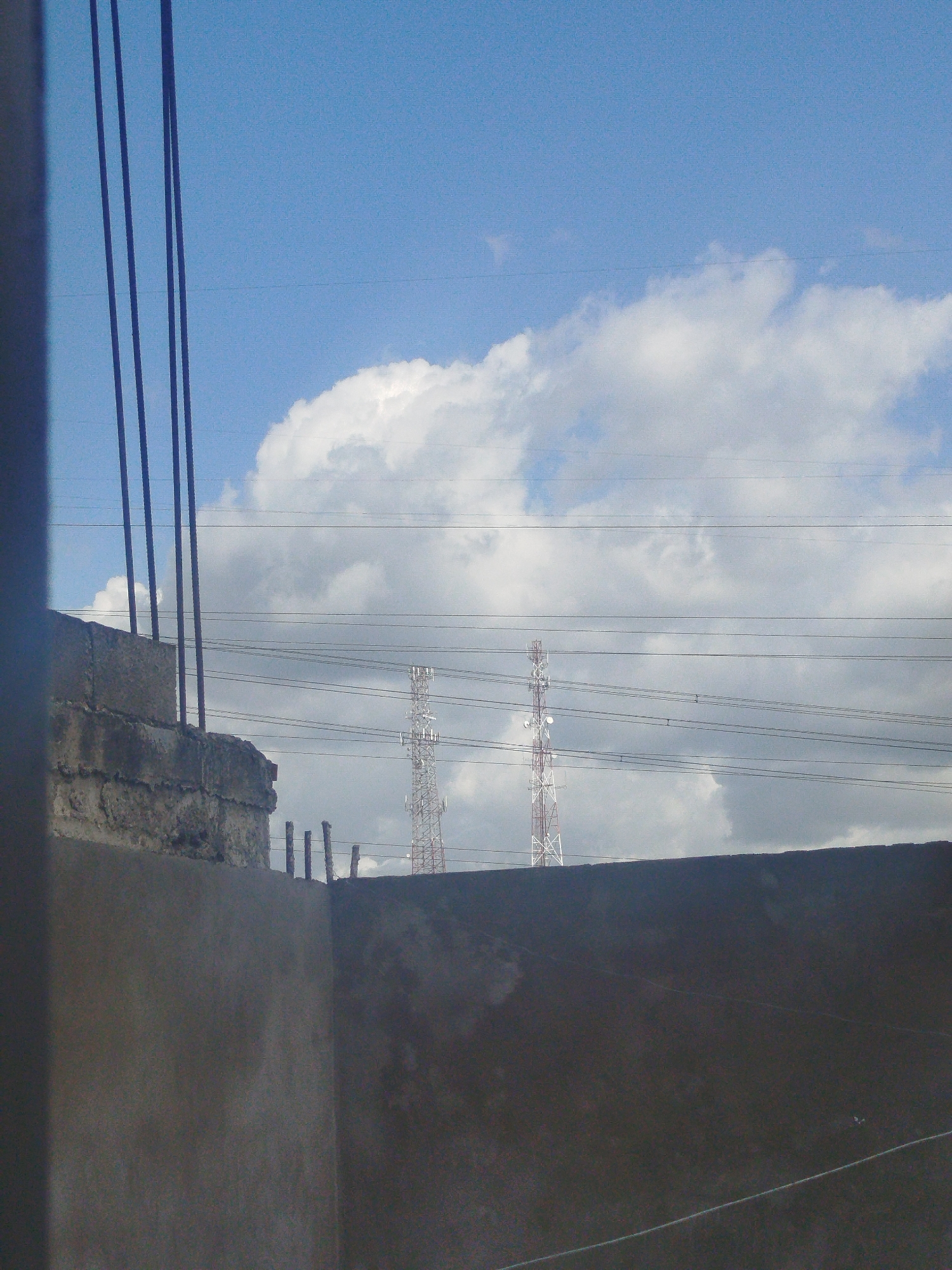The image, taken in Cúa, Venezuela, shows a partial view from behind a concrete structure. Visible over the structure are two lattice towers against a backdrop of a cloudy sky. Multiple overhead power lines are stretched across the sky. To the left of the image, rebar extends upwards from a concrete block.