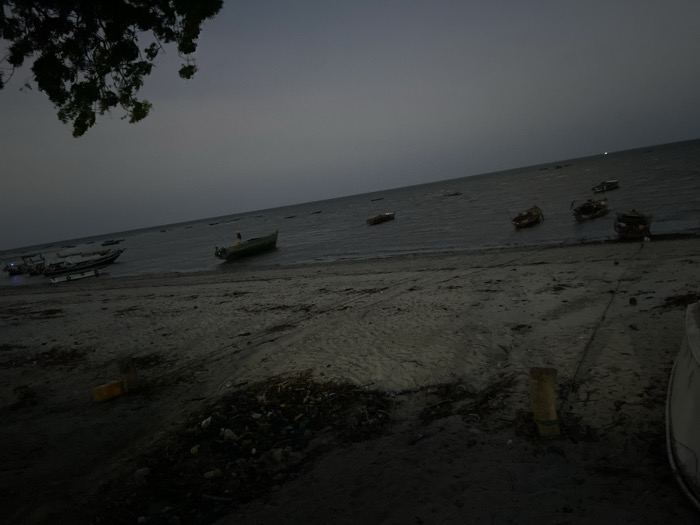 The image presents a dark, coastal scene at late dusk or early night, likely in Bagamoyo, Tanzania. The sky is uniformly dark gray and overcast, casting a somber light over the landscape.
In the foreground, a sandy and muddy beach occupies the lower half of the frame. Towards the bottom center, there is an accumulation of dark debris or trash, and to its left, a bright yellow bucket or container is visible on the sand. A thick wooden post stands upright in the sand to the right of the debris, with a rope extending towards the right edge of the frame. Part of a white boat hull is cropped at the far right.
Numerous traditional wooden boats are present across the scene. On the far left, one boat, partially beached, shows a faint blue light or reflection. Another boat rests closer to the water's edge towards the mid-left. Several more boats are afloat in the calm, dark water, particularly on the right side of the image where they appear clustered. The water itself is dark and still, reflecting the dim ambient light.
Dark, silhouetted tree branches with sparse leaves overhang the scene from the top left corner. The overall atmosphere is quiet and still, suggesting a peaceful end to the day in a working coastal community. No people are visible, and no discernible text appears anywhere in the image.