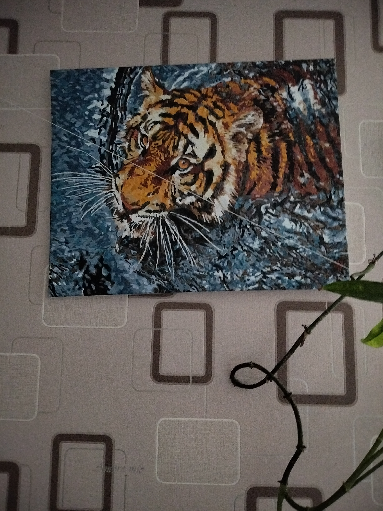 This image shows a painting of a tiger partially submerged in water, hanging on a wall. The painting features the tiger's head and shoulders, with its orange and black stripes clearly visible. The water appears dark and rippled. The wall behind the painting has a textured wallpaper with a repeating pattern of rounded rectangles in light and dark gray. A thin, dark vine with small nodes and a few green leaves winds across the lower right portion of the image, partially obscuring the wall and the bottom of the painting. The painting is positioned on the wall in a room. No specific location in Nizhnedonskoy, Russia can be identified from the image.