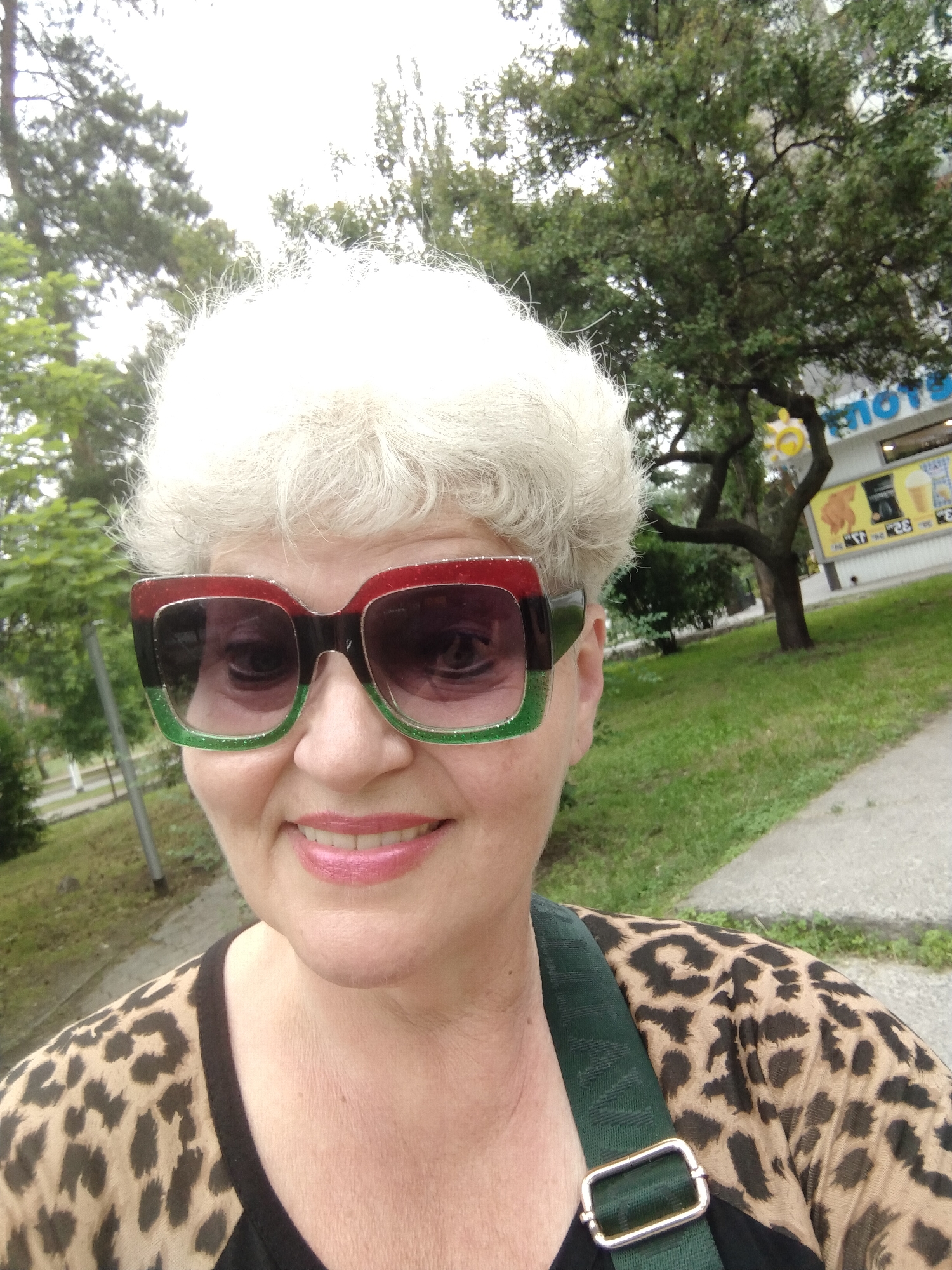 A woman with white, curly hair and large, square sunglasses with red and green frames smiles at the camera outdoors. She is wearing a leopard-print top. In the background, there are trees, green grass, a sidewalk, and a building with signage. The location is identified as Horishni Plavni, Ukraine.