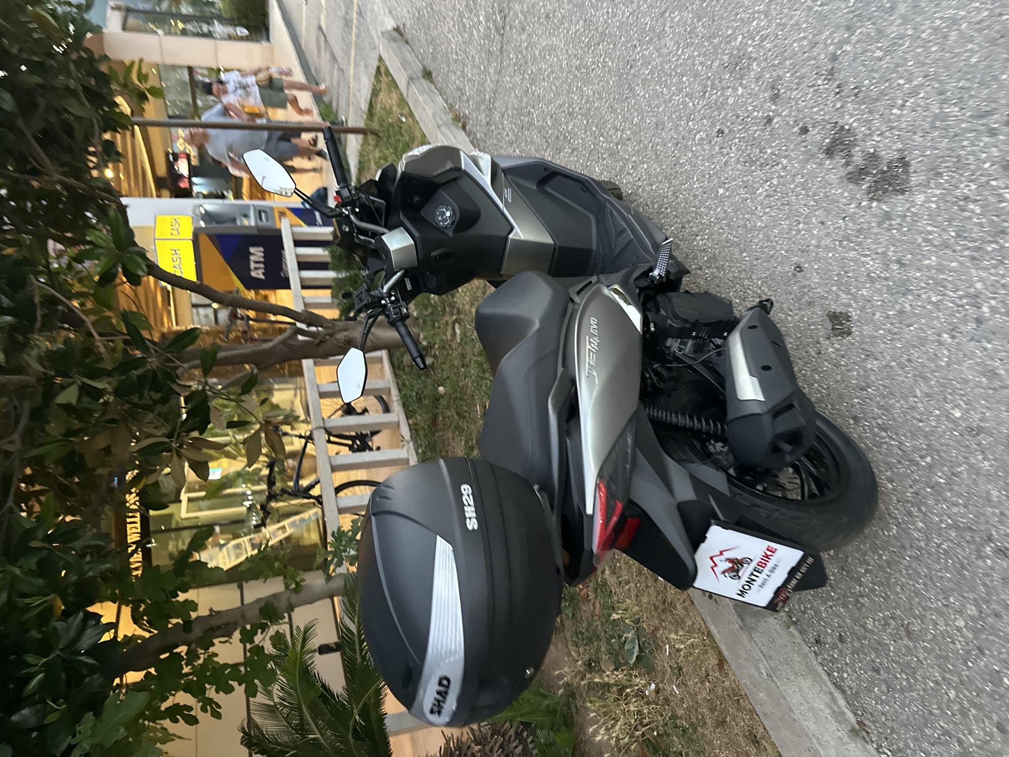 A parked grey and black scooter is in the foreground, facing right, with a black SHAD SH29 top box on the back. The scooter has "JET EVO" written on its side. The license plate holder features a "MONTEBIKE Rent A Bike" sticker with a red mountain logo. The scooter is parked on an asphalt surface next to a concrete curb and dry grass. Behind the scooter, a tree with green leaves overhangs the scene. In the background, there is a building with large windows, a yellow ATM machine, and people walking. The scene appears to be in an urban environment, possibly a street or pedestrian area in Budva, Montenegro, given the "MONTEBIKE" branding, a company that rents bikes and scooters in Montenegro.