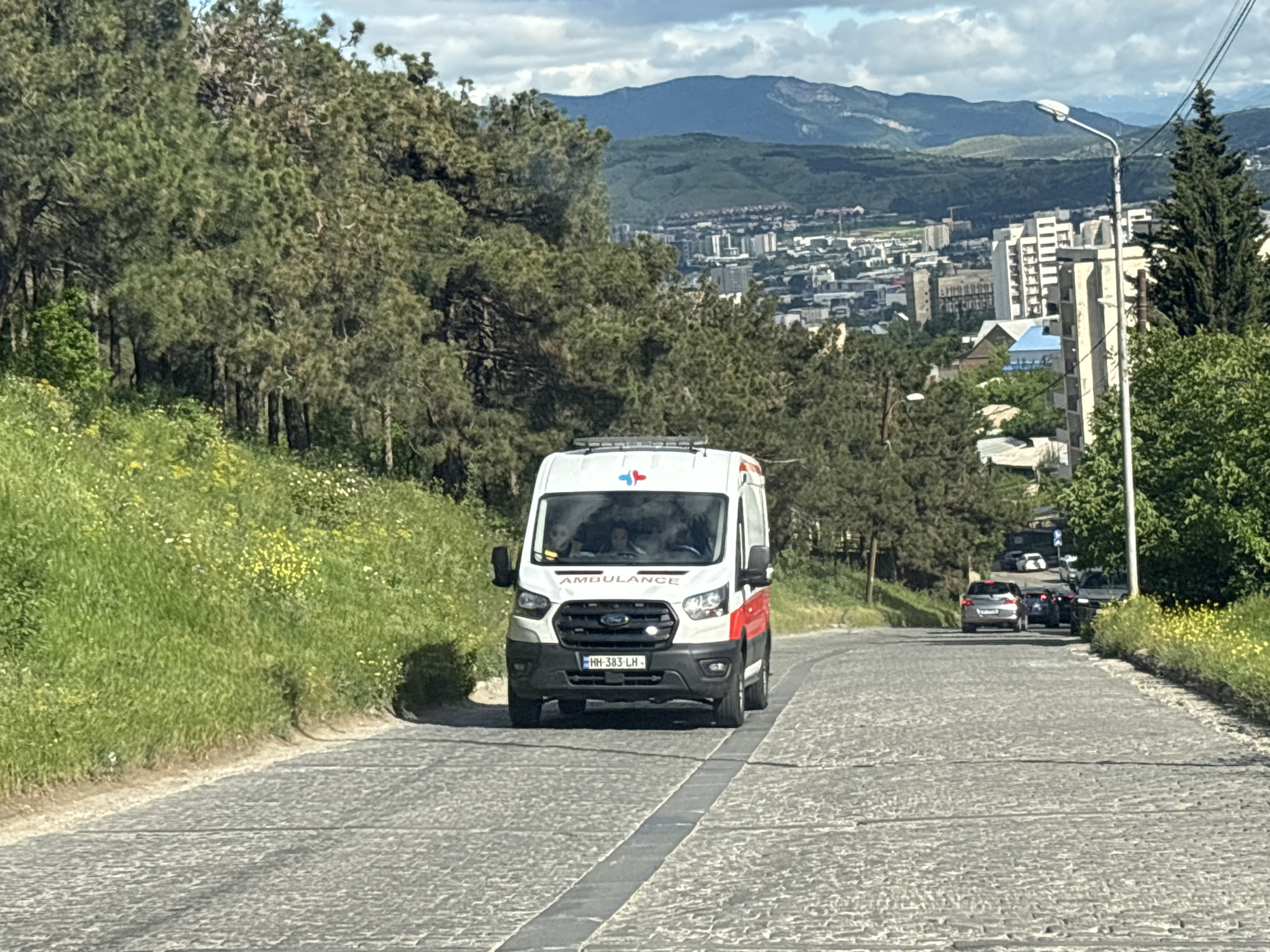 An ambulance is seen traveling up a cobblestone street in Tbilisi, Georgia. The ambulance is a white Ford Transit van with red and blue medical insignia. It has a license plate number "HH-383-LH" and the word "AMBULANCE" written on the front. The road is lined with green grass, trees, and yellow wildflowers on the left side and trees on the right. Further up the road, several cars are visible. In the background, there is a cityscape with buildings and mountains under a cloudy sky. A streetlight stands on the right side of the road.
