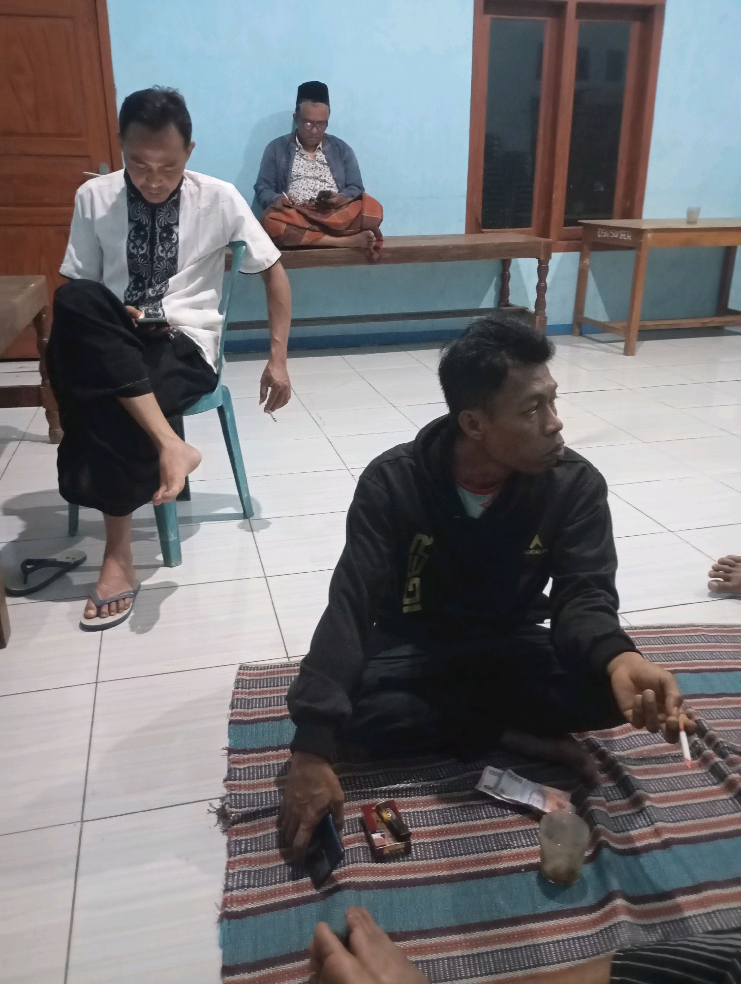 In Ketepung, Indonesia, three men are present in a dimly lit room. One man, dressed in a white shirt with black trim and black pants, sits on a blue chair, holding a lit cigarette and looking down at his phone. Another man, wearing a dark hooded sweatshirt, sits cross-legged on a striped mat on the floor. He is also holding a lit cigarette and looking to his right. A third man, in the background, sits on a wooden bench wearing a patterned shirt, a dark jacket, and a sarong, also looking at his phone. The room has light blue walls and a tiled floor. A wooden table and a bench are visible along the back wall, along with a wooden-framed window. On the mat in front of the man on the floor are several items, including a lighter, cash, and a glass with a beverage.
