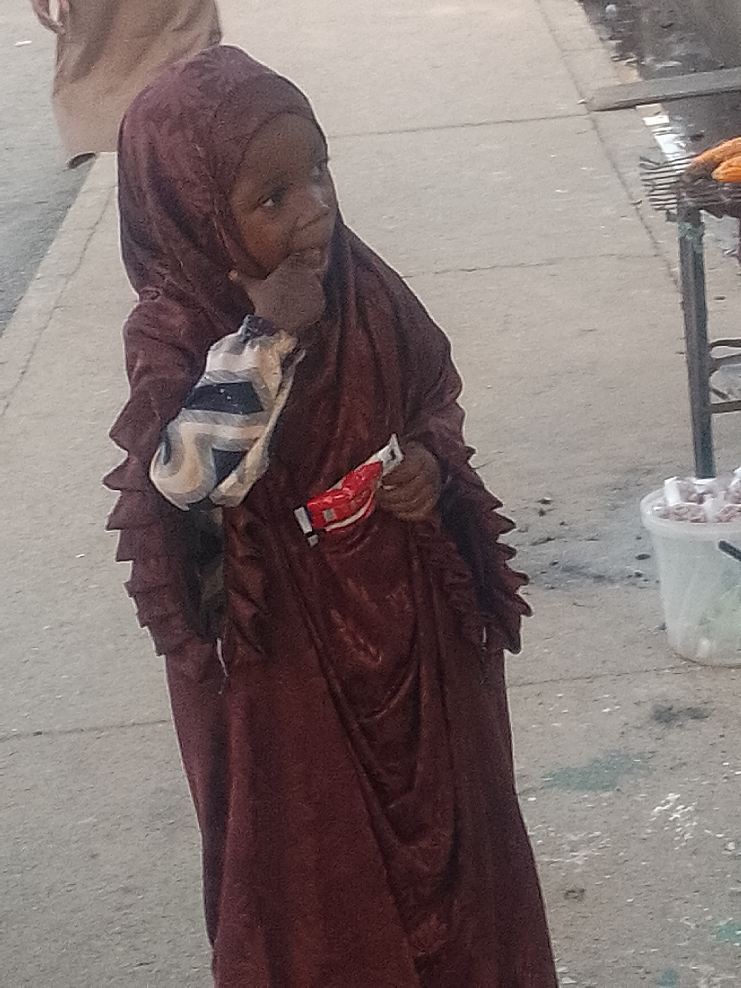 The image displays a young child standing on a light gray paved surface, consistent with a sidewalk, in Pantame, Nigeria. The child, with dark skin, is dressed in a full-length, dark maroon garment and a matching hijab. The garment features prominent ruffled or pleated trim along the sleeves. The child's right hand is positioned near her mouth, while her left hand holds a small, red and white packaged item. A separate, striped fabric in white, black, and beige is visible around her left arm. The child's head is turned, and she is looking towards the right side of the frame. In the upper left background, the lower torso of an adult, wearing light brown clothing, is partially visible and out of focus. To the right, a metal grill or rack holds several orange-yellow food items, suggesting a street vendor setup. Below the grill, a translucent white plastic bucket filled with numerous small, packaged items rests on the ground. The paved surface shows minor irregularities and scattered debris.