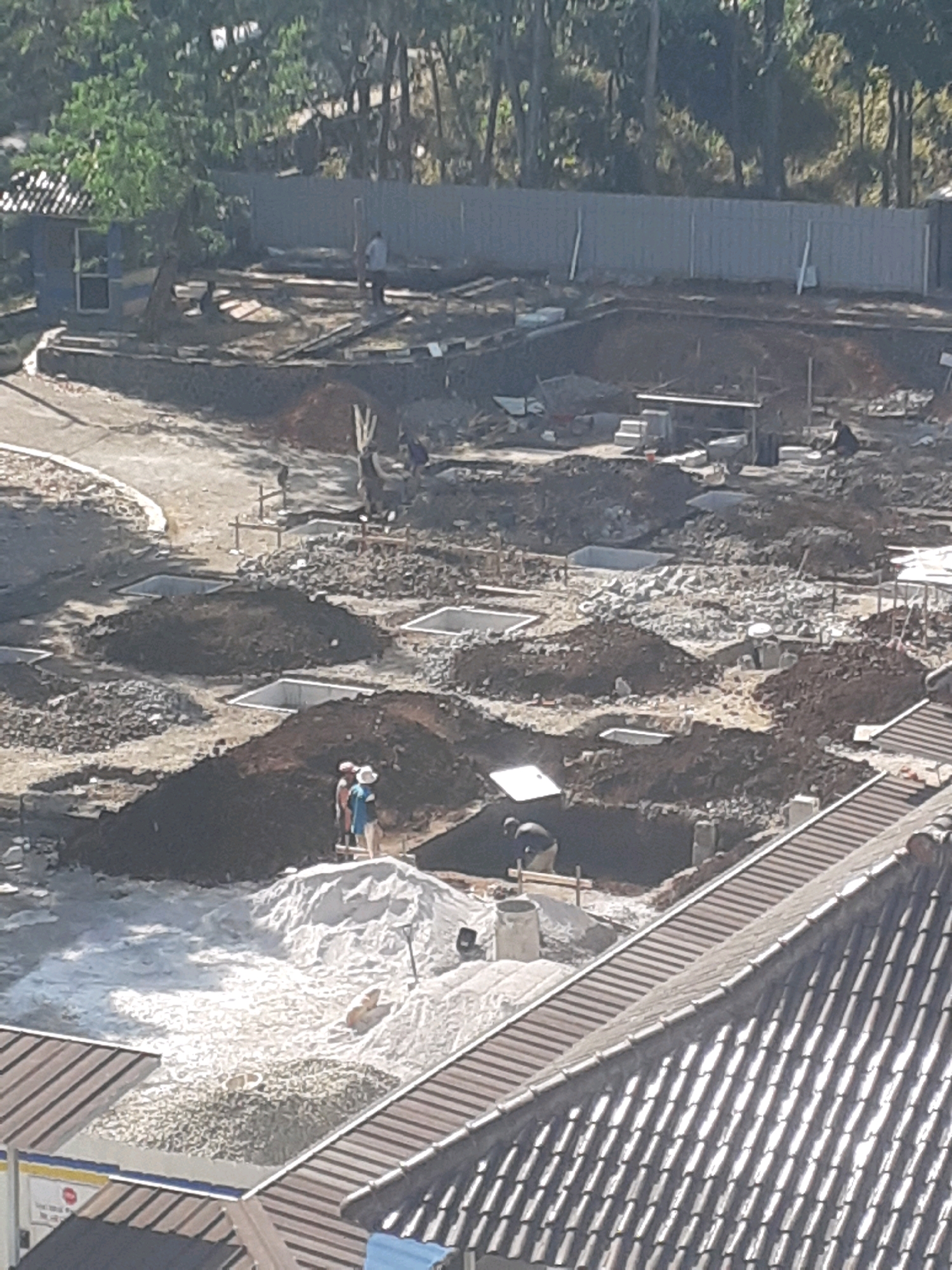 The image shows a construction site in Sumedang, Indonesia. There are several excavated pits, some of which are lined with concrete. Piles of dirt and gravel are scattered around the site. Several workers are visible, some wearing hard hats. One group of workers appears to be standing on a pile of white sand, possibly preparing to pour concrete. In the background, there is a fence and trees. A small building with a blue roof is also visible on the left side of the image. The scene suggests ongoing construction work, likely for building foundations or a similar structure.