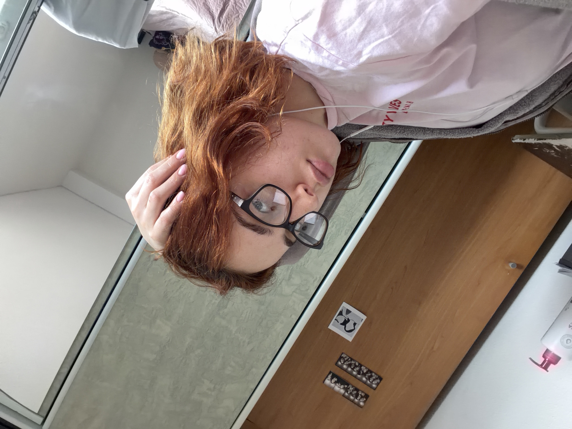 The image, taken in Strasbourg, France, depicts an indoor scene with a person lying down. The individual has reddish-brown, wavy hair and wears black-framed glasses. They are dressed in a light pink t-shirt, underneath which a grey garment (possibly a jacket or blanket) is visible. Wired earbuds are draped around their neck. Their right hand, with light pink nail polish, is gently placed near their temple, touching their hair. The person's expression is neutral, and their gaze is directed slightly downwards.
The background features a bright white wall or ceiling on the upper left, suggesting natural light, indicating it is daytime. A light grey, textured surface, possibly a wall or mirror, runs diagonally through the middle. To the right, a wooden surface, likely a desk or floor, displays a small black and white photo print and two strips of photo booth-style images, possibly of faces. A white appliance with a pink or red glow at its base is partially visible on the far right. Partial text "TY WES" and "RAW" is discernible on the pink t-shirt.