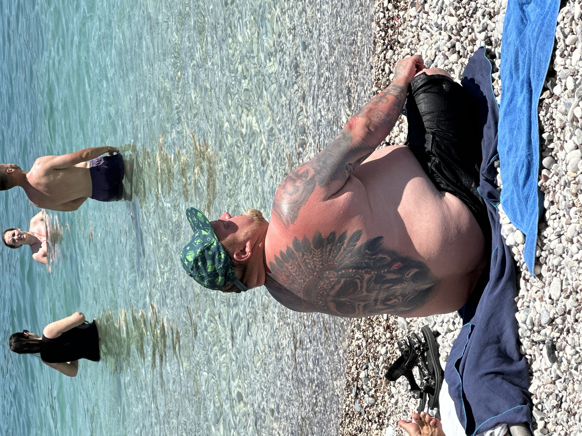 The image depicts a beach scene in Budva, Montenegro. In the foreground, a man with extensive back and arm tattoos, wearing a green patterned baseball cap and black swim trunks, is seated on a pebble beach, facing away from the camera. The water behind him is clear and shallow, with the seabed visible. Several other individuals are in the water. To the left of the seated man, a pair of black sandals rests on a dark blue towel. Further back, a woman in a black swimsuit stands in the water, and another woman with light-colored hair is partially submerged. In the mid-ground, a man in dark swim trunks stands in the water. The environment is sunny, with the water appearing calm.