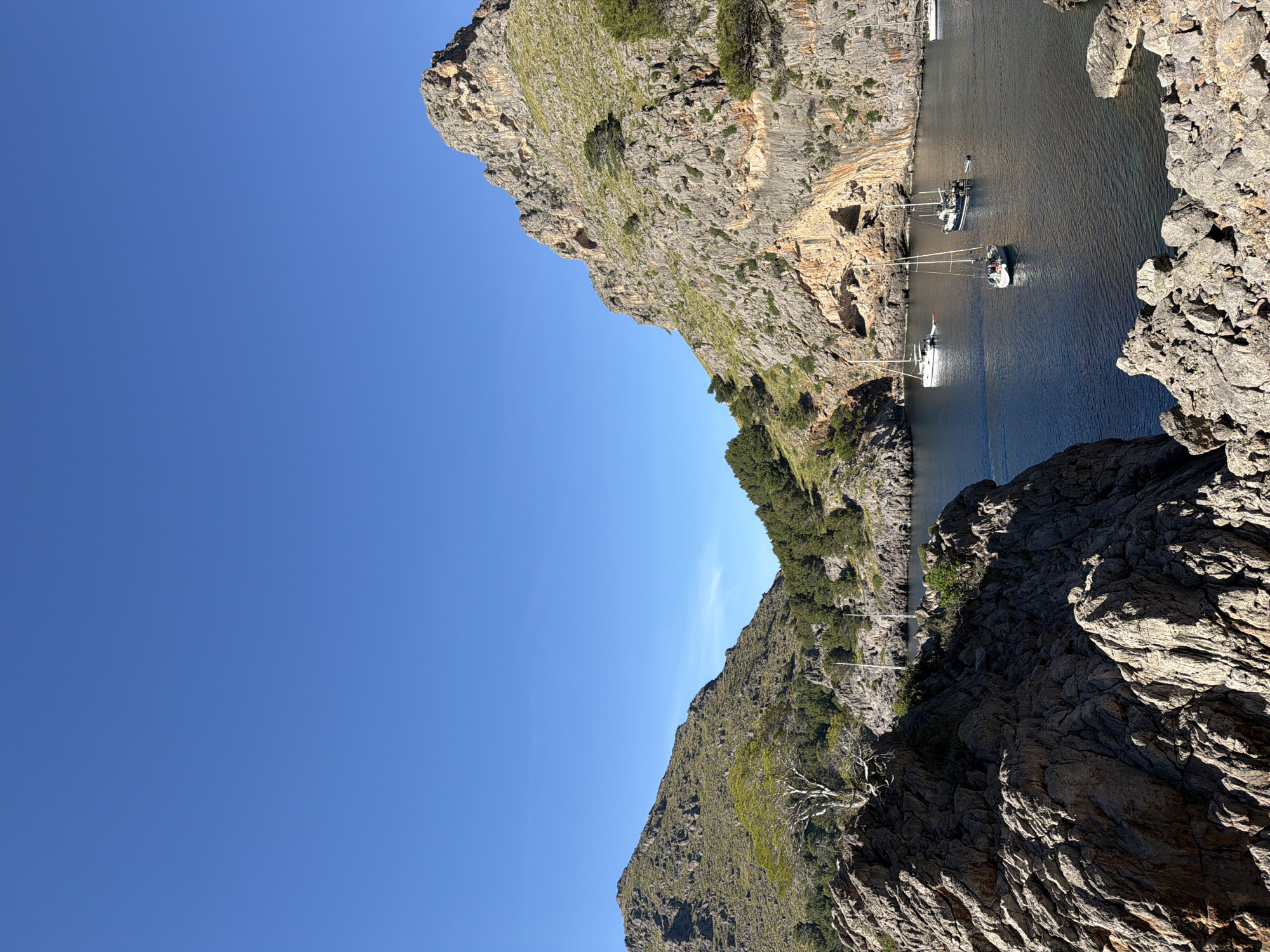 The image shows a view of Cala de Sa Calobra, a cove in Escorca, Spain. The scene is framed by rocky cliffs on both sides, with the blue sky filling the upper left portion of the image. The water in the cove is visible between the cliffs, where several boats are docked. The cliffs are rugged and textured, with some vegetation growing on them.