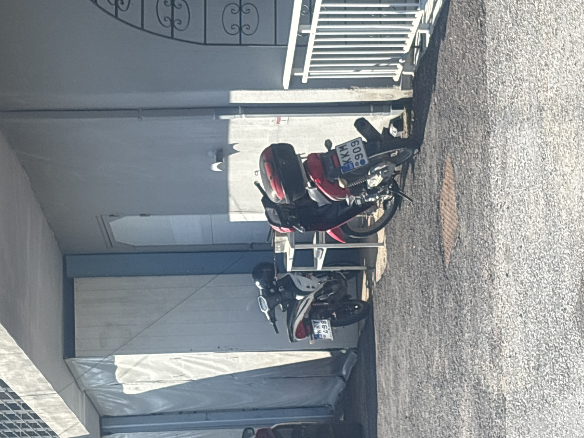 In the city of Kikidio, Greece, two motorcycles are parked on a gravel surface in front of a building. The motorcycle on the left is white and black and displays a license plate reading "KRY 498". The motorcycle on the right is red and black and has a license plate that reads "KKM 909". A white metal table is situated between the two motorcycles. The building behind the motorcycles has a light gray exterior and a blue trim around a doorway. A white metal fence is visible to the right of the motorcycles. The image appears to be taken during the daytime, with shadows indicating sunlight.