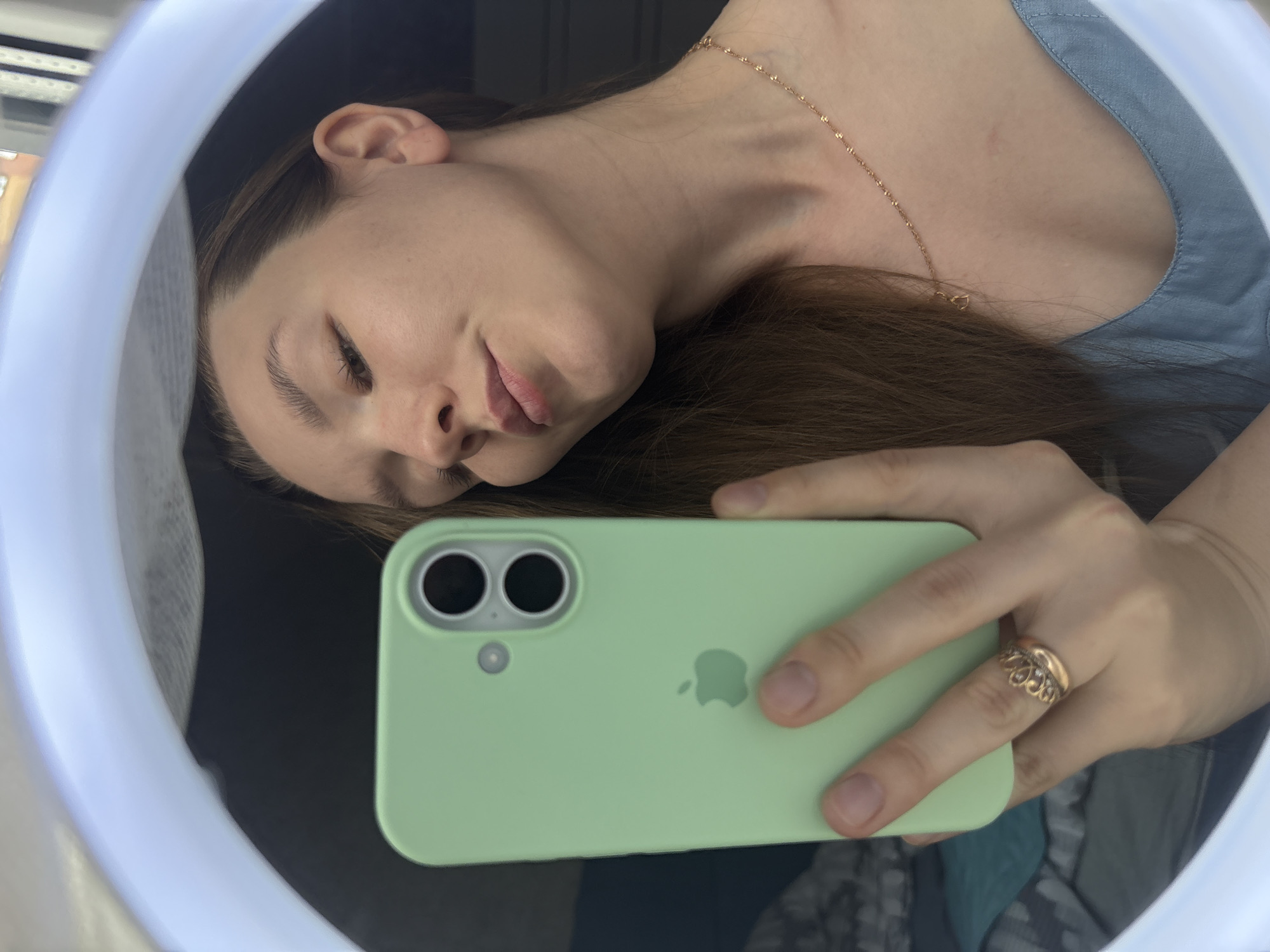 A young adult female is captured in a mirror selfie, taken in an indoor setting in the city of Izhevsk, Russia. The scene is illuminated by the white ring of a ring light that frames her reflection. She has long, straight, medium brown hair and light skin, with her lips slightly pursed as she looks down towards the phone. She wears a light blue, V-neck strap top or dress. Her right hand, adorned with a gold-toned crown-motif ring with small clear stones on the ring finger, holds a mint green iPhone with a visible Apple logo and a dual camera system on the back. A thin, gold-toned chain necklace with a small heart pendant is visible on her décolletage. The reflected background includes patterned fabric, a dark surface, and a partial view of a window with vertical elements, consistent with a domestic interior.