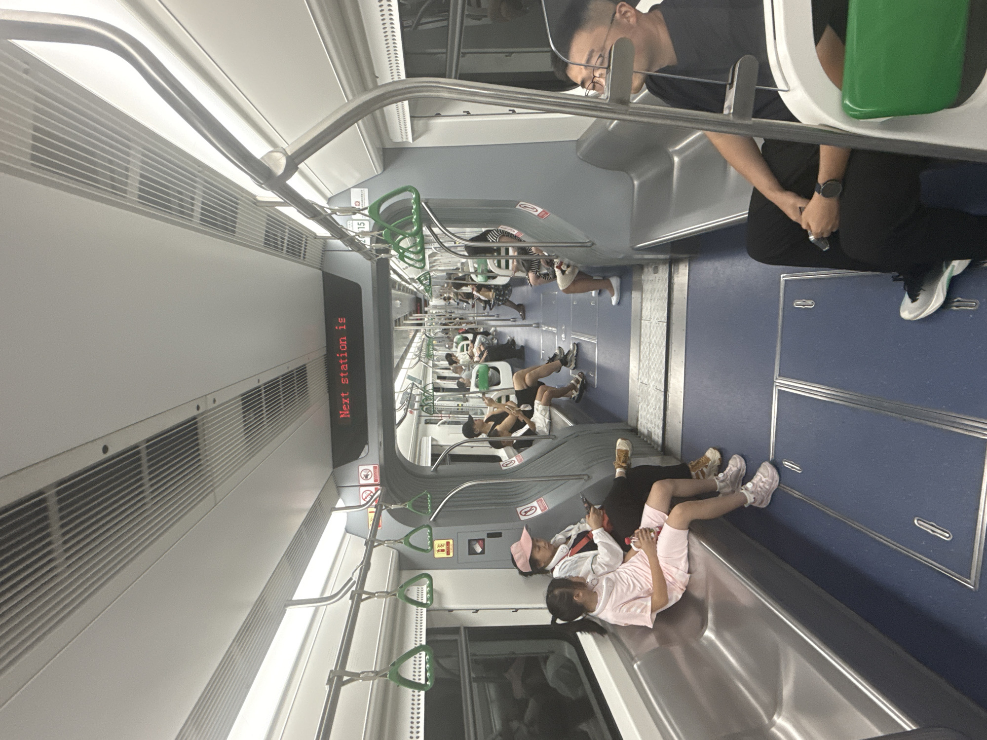 This is a photograph taken inside a subway car in Yuzhong District, China. The car is well-lit and appears to be modern, with rows of silver seats on either side and a blue floor with silver accents. Overhead, there are handrails and green triangular handles for passengers to hold onto. A digital display above the window reads "Next station is". Several passengers are seated, including a woman in a pink hat and a young girl in a pink outfit in the foreground. Other passengers are visible further down the car, some looking at their phones. The overall impression is one of everyday urban transit.