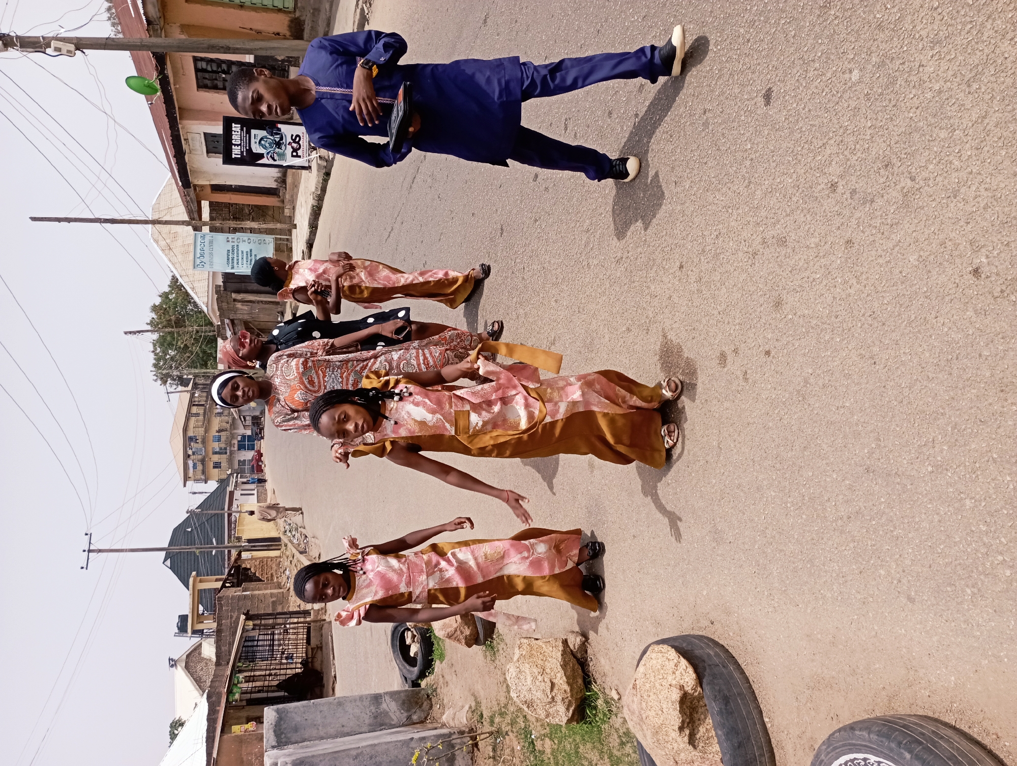 The scene depicts a group of people on a street in Jos, Nigeria. There are four girls and one boy. The girls are in a line, wearing matching dresses. The dresses are pink and gold with flowing designs. The boy is in the background wearing a blue shirt and pants, he is holding a black object. They appear to be posing for a photo. The street is paved. Behind the group is a typical African street scene with a building, wires, and signs on posts. There are tires and rock placed on the side of the road. The lighting suggests it is daytime.