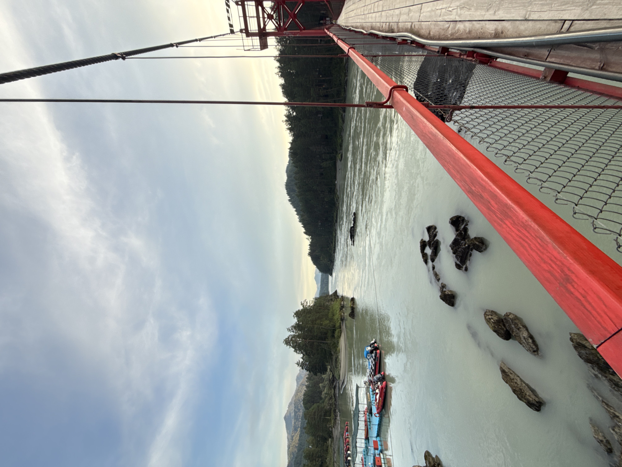 The image shows a view from a suspension bridge in the city of Barangol, Russia. The bridge has a red railing and a wooden walkway. A river flows below the bridge, with boats docked on the left side. Trees line the banks of the river, and mountains are visible in the distance. The sky is partly cloudy, with the sun setting in the background, casting a warm glow on the scene.