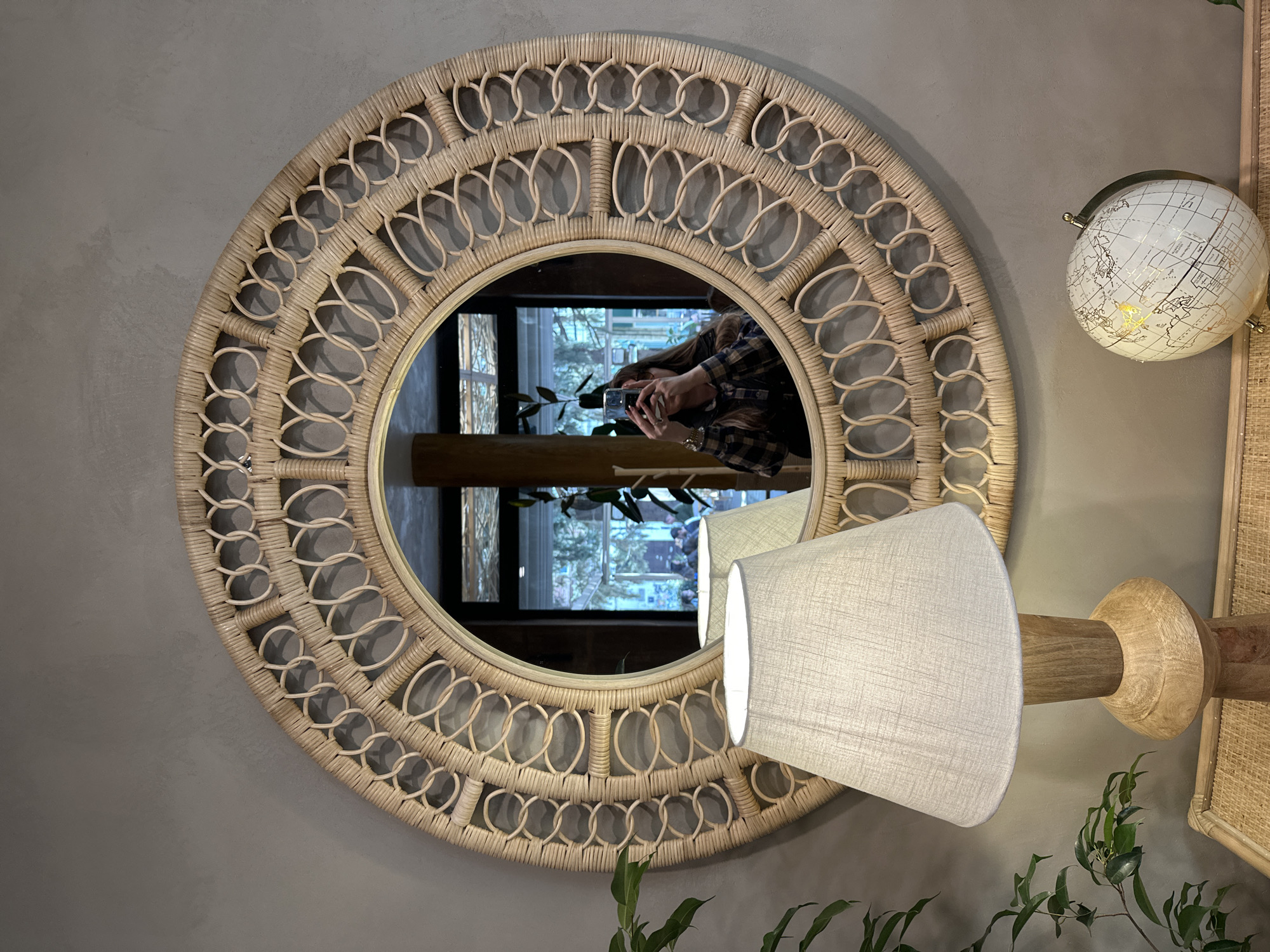 A round wicker mirror hangs on a gray wall in what appears to be an interior in Tbilisi, Georgia. The mirror reflects a woman in a plaid shirt holding a smartphone, capturing the image of herself. Outside the window reflected in the mirror, there are trees and a building. To the right of the mirror, there is a globe and a lamp with a beige lampshade on a wooden stand. Green leafy plants are present near the lamp. The items suggest a well-lit and decorated interior space.